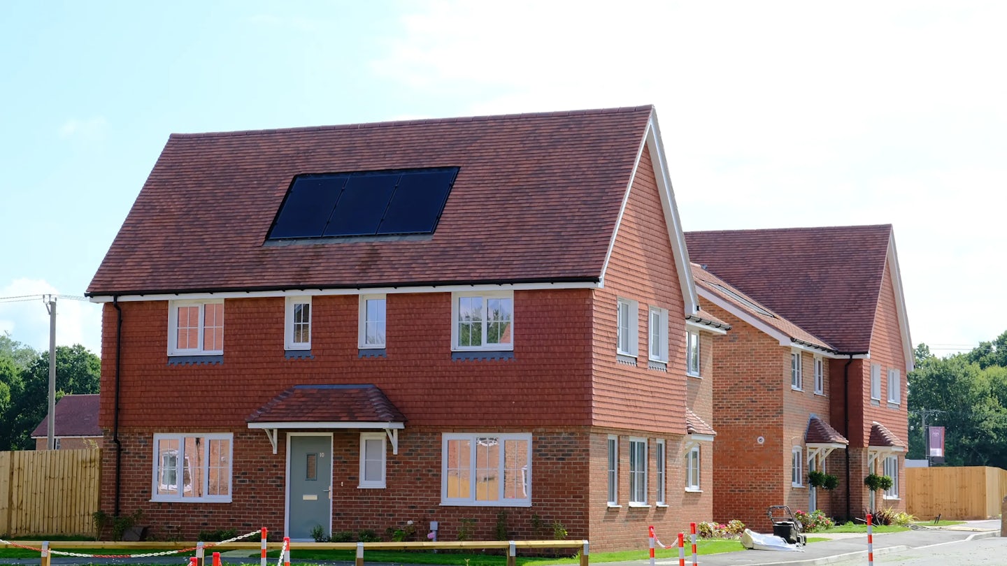 Newly built brick house with a red-tiled roof and a single solar panel installed. A similar home is visible nearby, and the area is partially fenced off with red-and-white tape, indicating ongoing construction or landscaping. The sky is partly cloudy, adding a sense of openness to the residential setting.