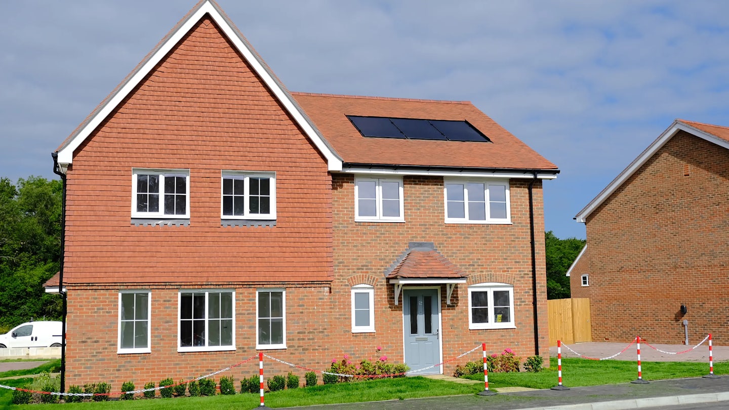 Modern two-story house with a red-tiled roof and brick exterior, featuring solar panels on the right side of the roof. A grey front door with a small canopy sits below evenly spaced windows. A white van is parked nearby, and the surrounding area includes grass, bushes, and red-and-white safety tape marking off part of the pavement. Another house is visible in the background under a partly cloudy sky.