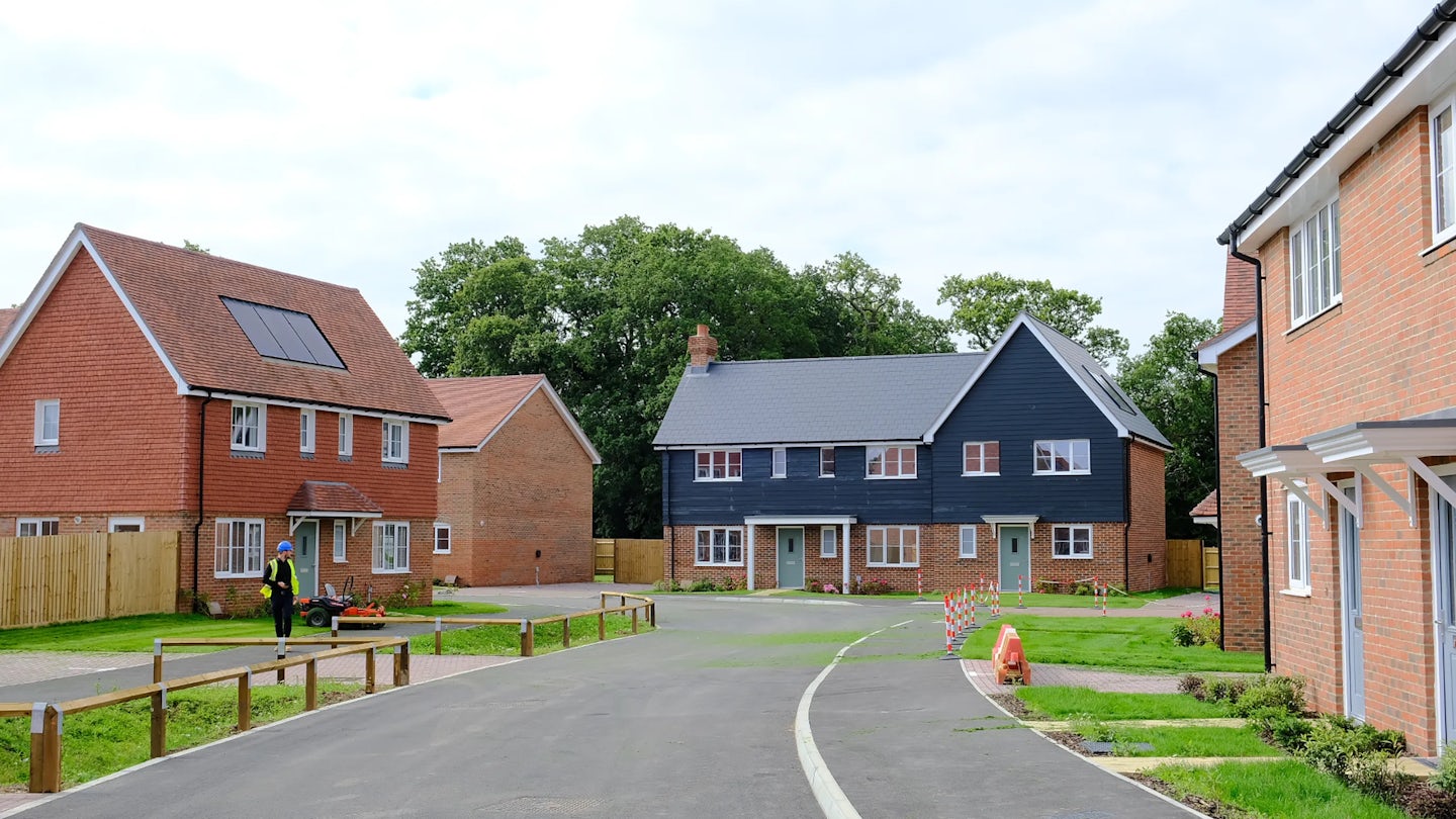 New-build residential street with three modern homes featuring solar panels, landscaped lawns, and tidy pavements. One person in a high-visibility jacket works near the grass. Facades vary—brick, dark blue cladding, and mixed materials—highlighting architectural contrast under a partly cloudy sky.
