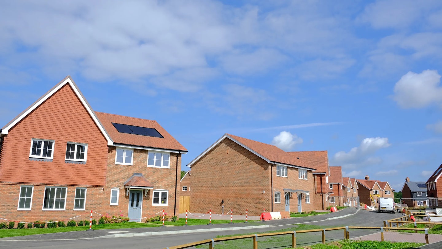 Newly built brick houses with red-tiled roofs in a tidy residential area under a bright, partly cloudy sky. One house has solar panels installed on its roof. Green lawns, road barriers, and traffic cones suggest ongoing landscaping or finishing work. Would you like a version that leans more into the sustainability message or highlights the sense of renewal and modern development? Happy to refine it with you, Aidan.