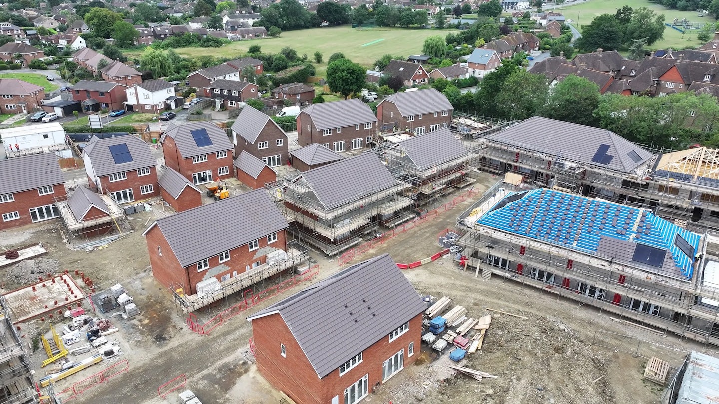 Aerial view of a suburban construction site with newly built homes in neat rows and several under active development. Some rooftops feature solar panels, while scaffolding and construction vehicles mark the progress of ongoing builds. The site borders existing homes and green fields, creating a contrast between established and emerging residential zones. A light overcast sky provides soft, even lighting over the expansive housing development.