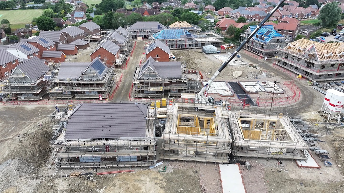 Residential construction site with rows of partially built homes, some with completed roofs and others in framing stages. A tall crane stands at the center, surrounded by construction materials, scaffolding, and vehicles. The site borders a neighborhood of finished houses, highlighting phased development and growth. Overcast sky adds neutral lighting to the busy, transitional environment.
