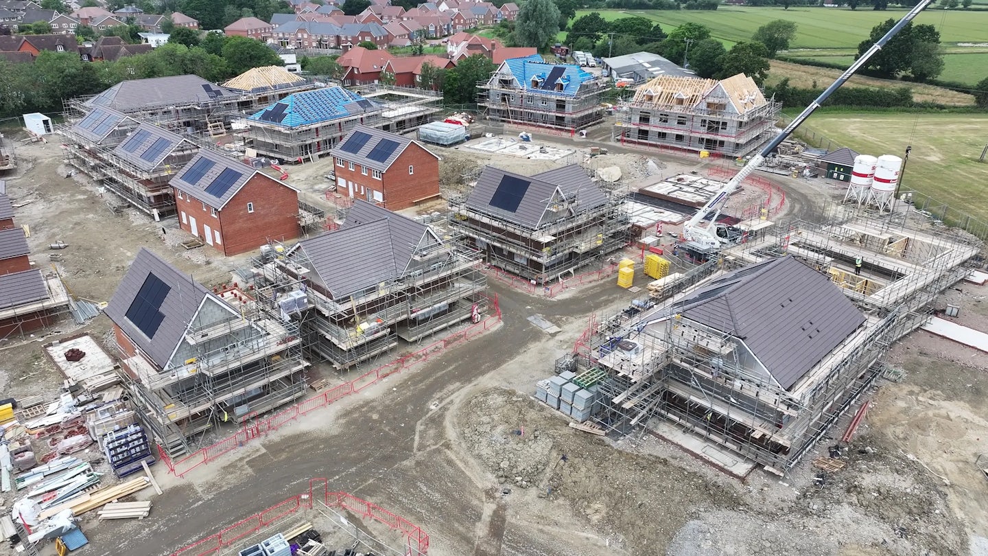 Wide-angle view of a residential construction site with multiple homes in various stages of development. Some rooftops feature solar panels, while others have scaffolding and exposed framing. Construction vehicles and workers are visible across the site, which is bordered by completed homes and open green fields. The structured layout and sustainable elements reflect eco-conscious growth and planned community expansion under a lightly clouded sky.