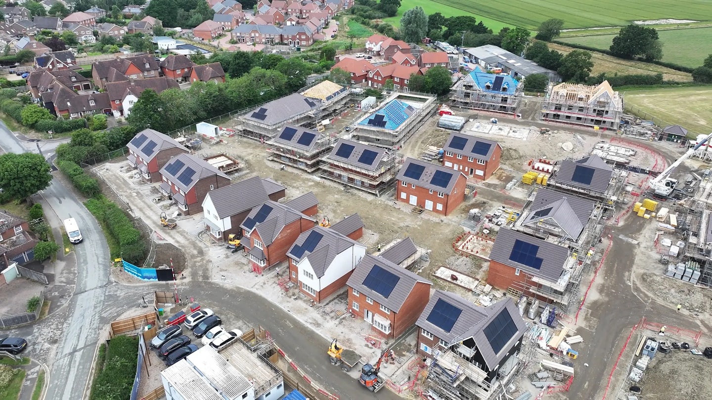 Aerial view of a large residential construction site featuring rows of new-build homes in various stages of completion. Many rooftops display installed solar panels, highlighting a focus on renewable energy. Construction vehicles and materials are scattered across the site, which is bordered by established neighborhoods and open green fields. The grid-like layout and surrounding greenery convey a sense of organized expansion and eco-conscious development.