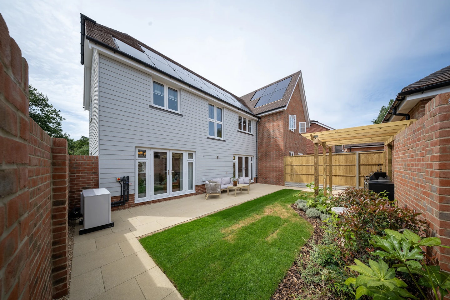Rear view of a modern suburban home featuring white siding, large windows, and rooftop solar panels. French doors open onto a landscaped garden with a lush lawn, paved patio, and outdoor seating area. A wooden pergola stands beside a brick wall, offering shade and architectural interest. Decorative planting beds frame the space, combining sustainability and comfort in a well-maintained backyard designed for relaxation and eco-conscious living.