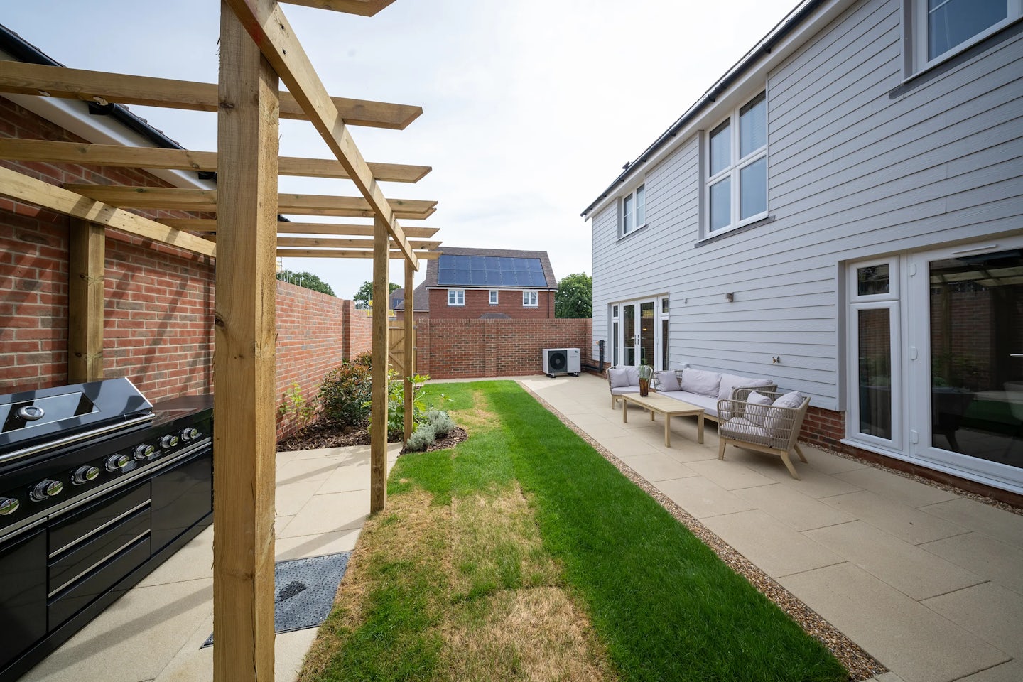 Modern backyard with a pergola sheltering a black barbecue grill on the left, bordered by paved walkways and a narrow strip of lawn running through the center. Outdoor seating with a sofa and chairs is arranged on the right, creating a relaxed social area. In the background, a neighboring building displays rooftop solar panels, reinforcing eco-conscious living. The space feels tidy, intentional, and designed for outdoor enjoyment.
