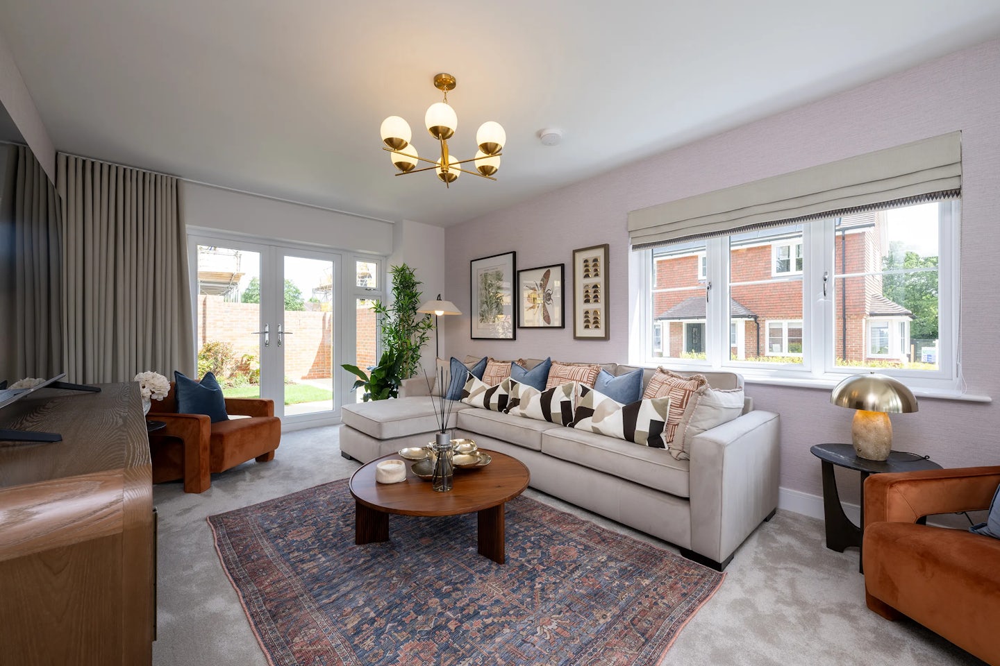 Bright living room featuring a large white sectional sofa with colorful patterned cushions, paired with a round wooden coffee table atop a geometric rug. Two brown armchairs sit opposite each other, framing the cozy setup. A wooden sideboard holds a flat-screen TV beneath framed artwork, while French doors and large windows invite natural light into the airy space. A chandelier and table lamp blend classic and modern elements, creating a warm, inviting atmosphere.