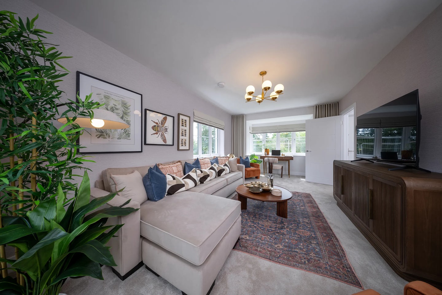 Elegant living room with a large beige sectional sofa, patterned rug, and wooden coffee table topped with a tray and decorative items. A flat-screen TV sits on a wooden console, while framed botanical prints adorn the neutral walls. A leafy potted plant adds greenery near the seating area. Natural light floods in through windows behind a desk and chair, forming a casual workspace. A modern chandelier hangs overhead, reinforcing the room’s cozy yet sophisticated atmosphere.