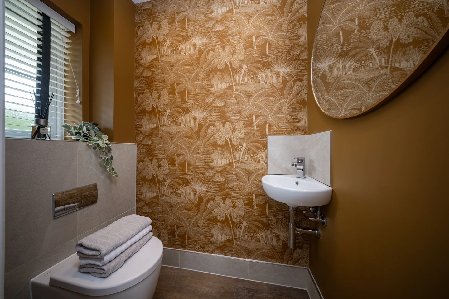 Compact, modern bathroom featuring a wall-mounted sink with a chrome faucet beneath a circular mirror. To the left, a white toilet is topped with neatly folded towels. The walls are decorated with tropical-themed wallpaper in warm brown and beige tones, and a window with white blinds allows natural light into the space. The cozy layout and artistic styling create a balanced blend of function and design flair. Would you like to adjust tone—say, for retrofit messaging, editorial storytelling, or platform-specific styling? I’m happy to dial in focus or texture.