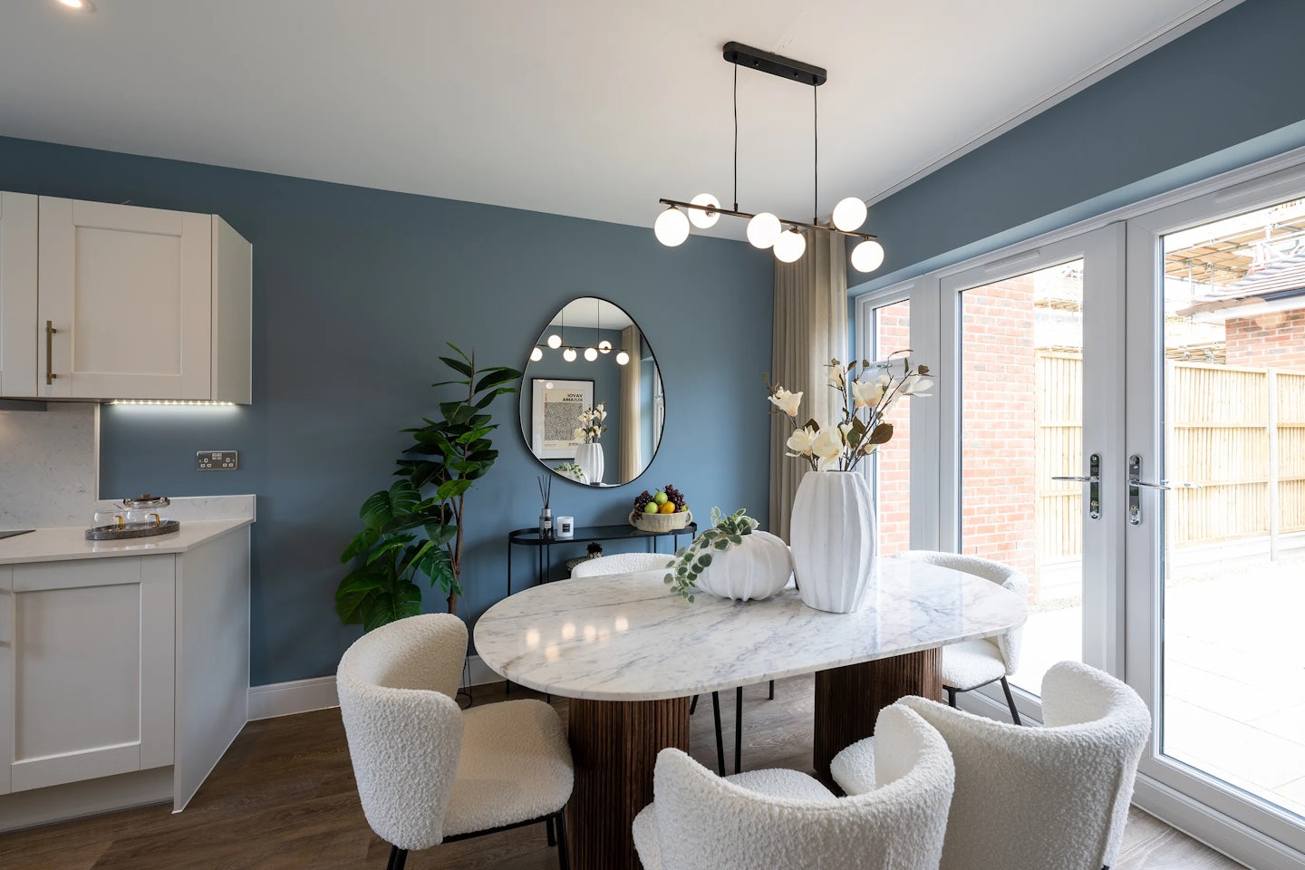 Stylish dining area featuring a round marble table surrounded by five upholstered chairs. A large white vase with flowers and a decorative white pumpkin sit at the center, under a chandelier with spherical lights. Behind the table, a deep blue accent wall holds a circular mirror and a small console with plants, adding warmth and visual interest. To the right, large glass doors open to an outdoor space, while part of the white kitchen is visible to the left, completing the clean and modern ambiance.