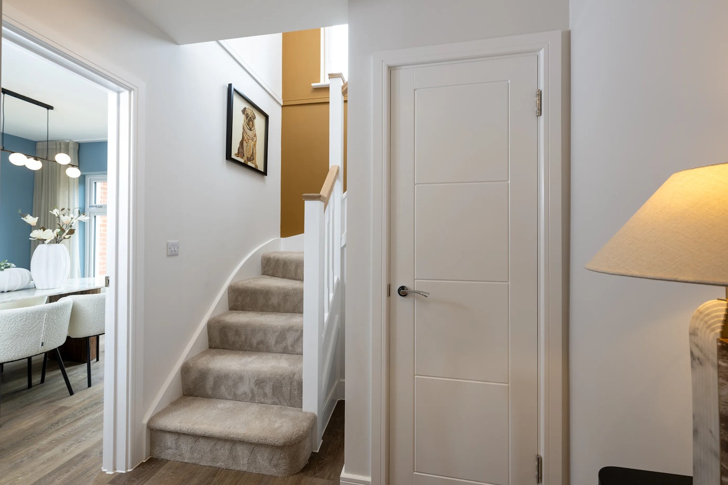 Interior view of a home featuring a carpeted staircase with white bannisters leading to an upper floor. A mustard yellow accent wall adds warmth above the stairs, which are framed by white walls. To the left, an open doorway reveals a dining area with a wood floor, floral centerpiece in a tall white vase, and surrounding chairs. A lamp with a beige shade sits on the right, beside a closed white door. A framed picture of a dog hangs above the staircase, adding a touch of character to the softly lit, family-friendly space.