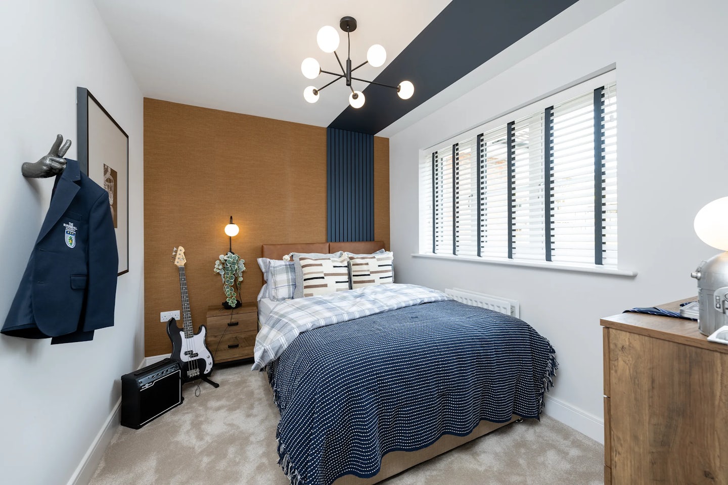 Stylish bedroom with a blue-and-white checkered duvet, layered pillows, and a wooden nightstand holding a lamp and flowers. A guitar and amplifier rest on the floor, adding a personal, musical touch. A blue blazer with a crest hangs on the wall above brown and blue decorative panels. Overhead, a modern chandelier with exposed bulbs provides ambient light.