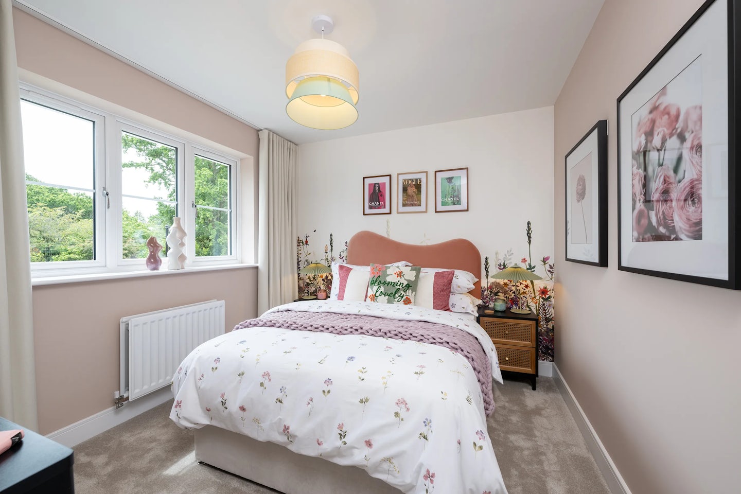Bedroom with a double bed dressed in a floral-patterned duvet and assorted pillows, including one with the word “LOVE” in bold text. Above the bed hang three framed prints, adding decorative charm. To the right, a wooden nightstand holds flowers and personal items, while a window on the left offers views of greenery and natural light. A modern ceiling light and neutral beige walls complete the serene, lived-in atmosphere.