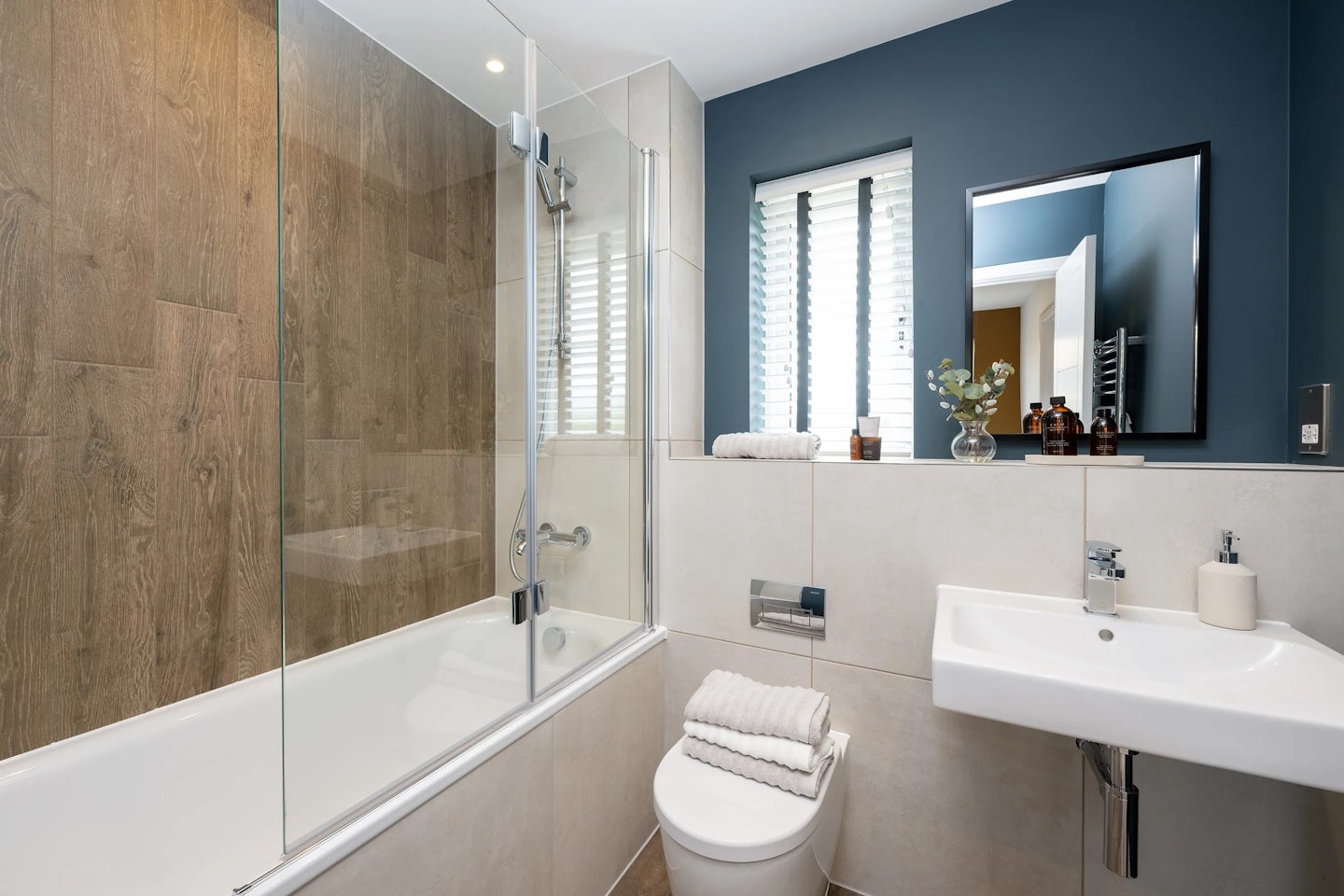 Contemporary bathroom featuring a bathtub-shower combo with a wood-textured backsplash and glass partition on the left. A toilet sits beside the tub, topped with folded white towels and backed by a small shelf with decorative items, including a vase of flowers. To the right, a white sink with a chrome faucet rests against a dark blue accent wall, with a mirror above and two narrow windows with white blinds letting in soft daylight. The space is tidy and thoughtfully styled, combining warmth, functionality, and modern design.