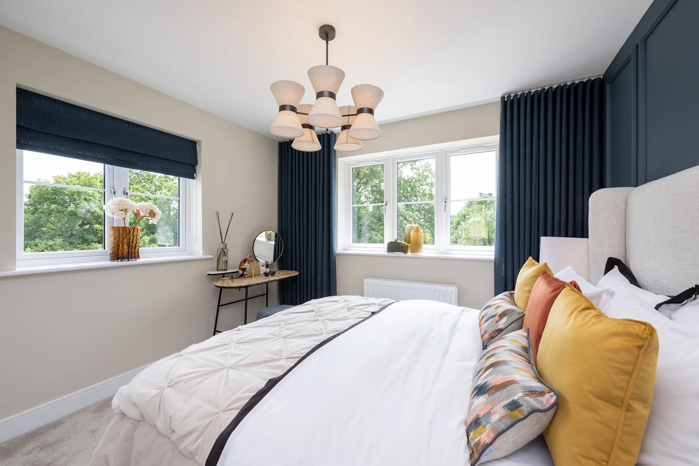 Stylish bedroom featuring a white bed with a mix of yellow, orange, and patterned cushions under a multi-arm chandelier. A large window with dark blue curtains allows soft daylight to illuminate a white side table adorned with decorative items, including a vase of flowers and a tabletop mirror. The walls are painted in a neutral tone, enhancing the room’s cozy and refined atmosphere. Want to shape the tone toward wellness, lifestyle branding, or retrofit storytelling? I can pivot the texture and detail focus accordingly.