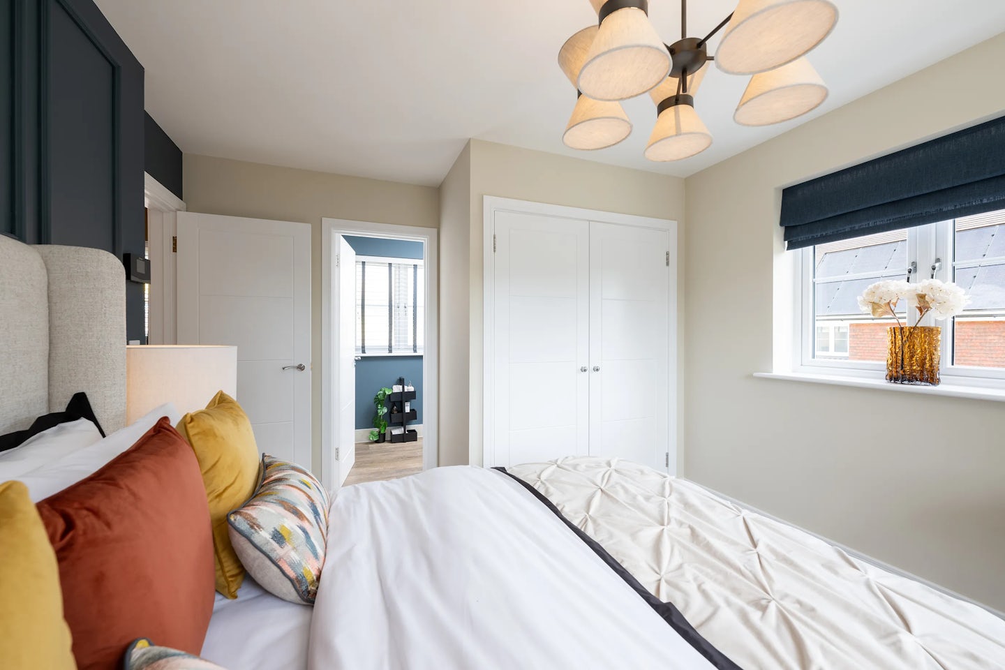 Modern bedroom with a neatly made bed dressed in white linens and accented by mustard yellow, burnt orange, and patterned pillows. A bedside lamp sits on the left, and a five-light chandelier hangs overhead. To the right, a window with a dark blue blind lets in soft natural light, with a vase of flowers on the sill. The room opens into a hallway through a white door, revealing a glimpse of another space with a blue wall and window. The cozy styling and warm color palette evoke comfort and thoughtful interior design.