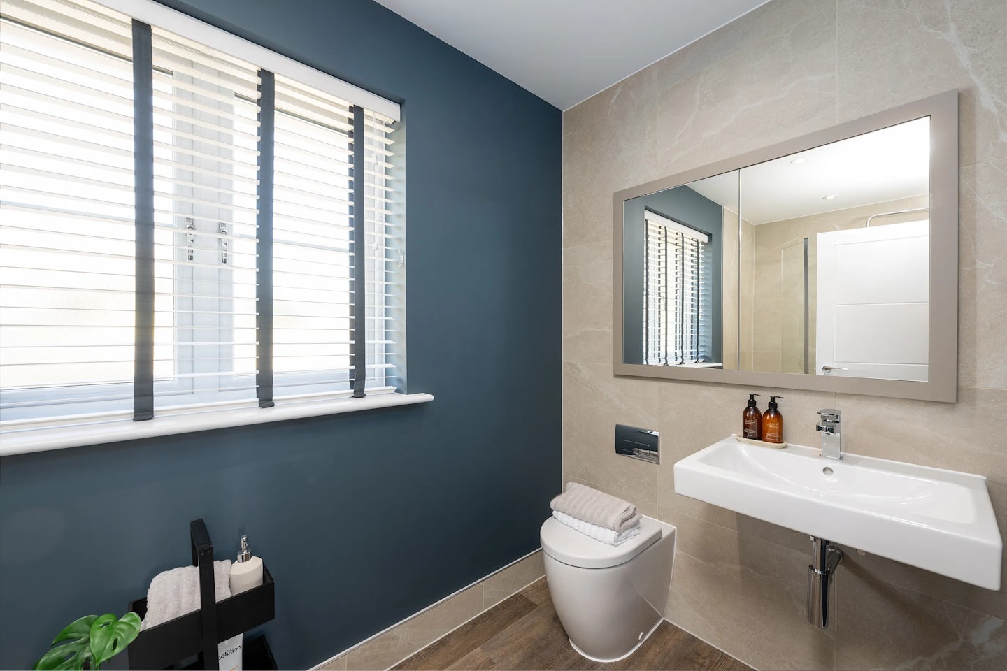 Modern bathroom featuring a white toilet, a matching sink with chrome faucet, and a large rectangular mirror above. The back wall is tiled in beige, contrasting with a dark blue side wall that includes a window with white blinds. Below the window, a black storage unit holds a plant and toiletries, adding a homely touch. Two soap or lotion bottles sit atop the sink, and the wood-textured floor enhances the room’s clean, contemporary look.
