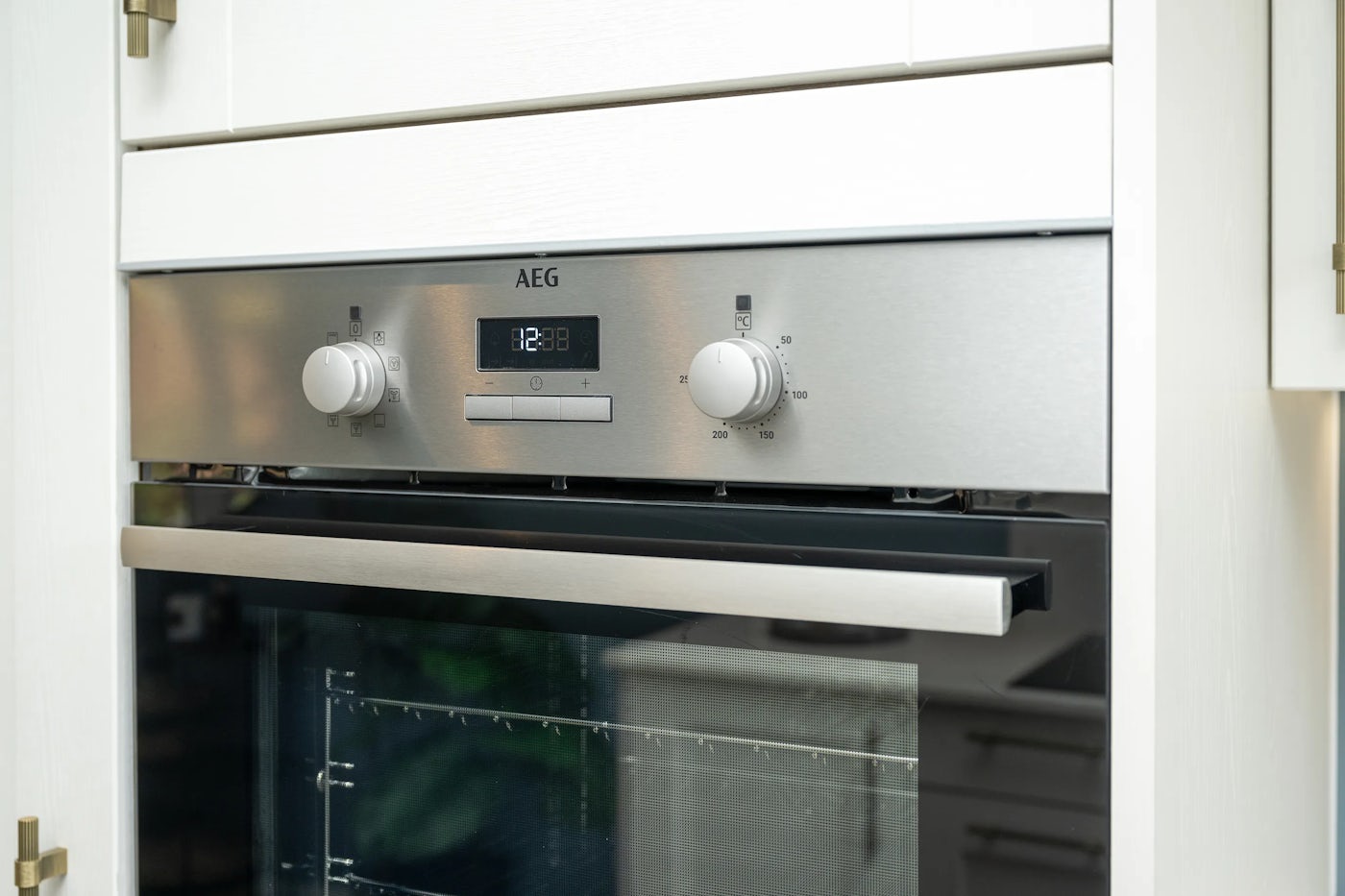 Built-in stainless steel oven with two control knobs and a central digital display showing the time “12:38.” The left knob appears to set oven functions, while the right adjusts temperature. The oven door is slightly ajar, revealing a wire rack inside. Branding “AEG” is visible above the display, underscoring sleek, modern design.