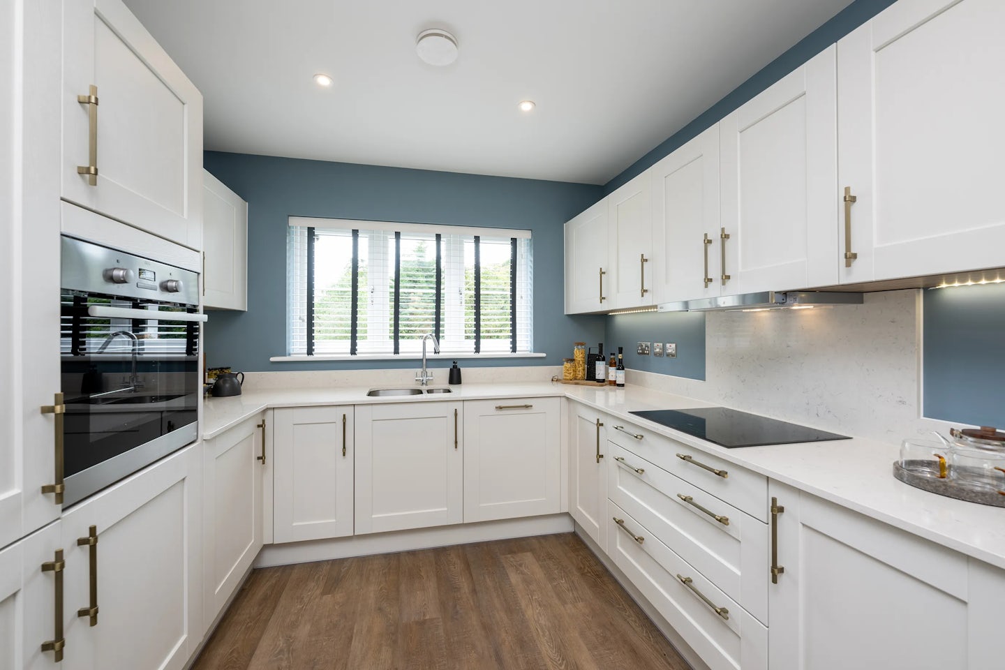 Modern kitchen featuring white cabinetry with brass handles, white countertops, and a light-toned backsplash. An induction cooktop sits beside a built-in oven, while the sink is positioned under a window with vertical blinds. The walls are painted deep blue, contrasting with the warm wood floor. Recessed ceiling lights provide ample illumination, and a tray with bottles adds a subtle styling touch.