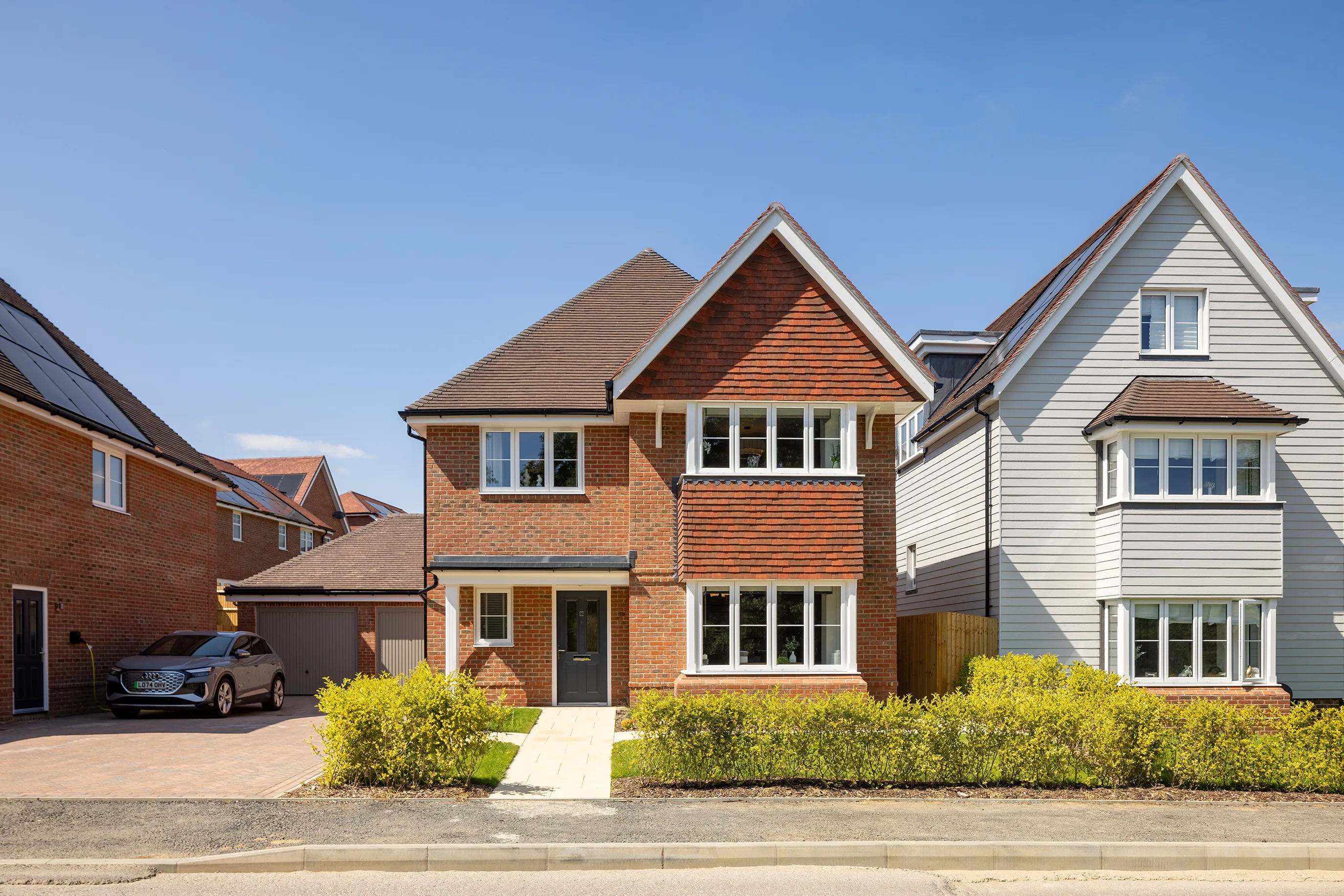 A two-story suburban house with a red brick exterior, pitched roof, and multiple white-framed windows, including a prominent bay window on the ground floor. The home sits beside a paved driveway leading to a garage, with a parked car partially visible. Flanked by neighboring houses with contrasting facades—one white and one brick—the scene features a neatly landscaped front yard with shrubs and a walkway to the entrance. Bright sunlight and a clear blue sky evoke a cheerful, residential atmosphere.