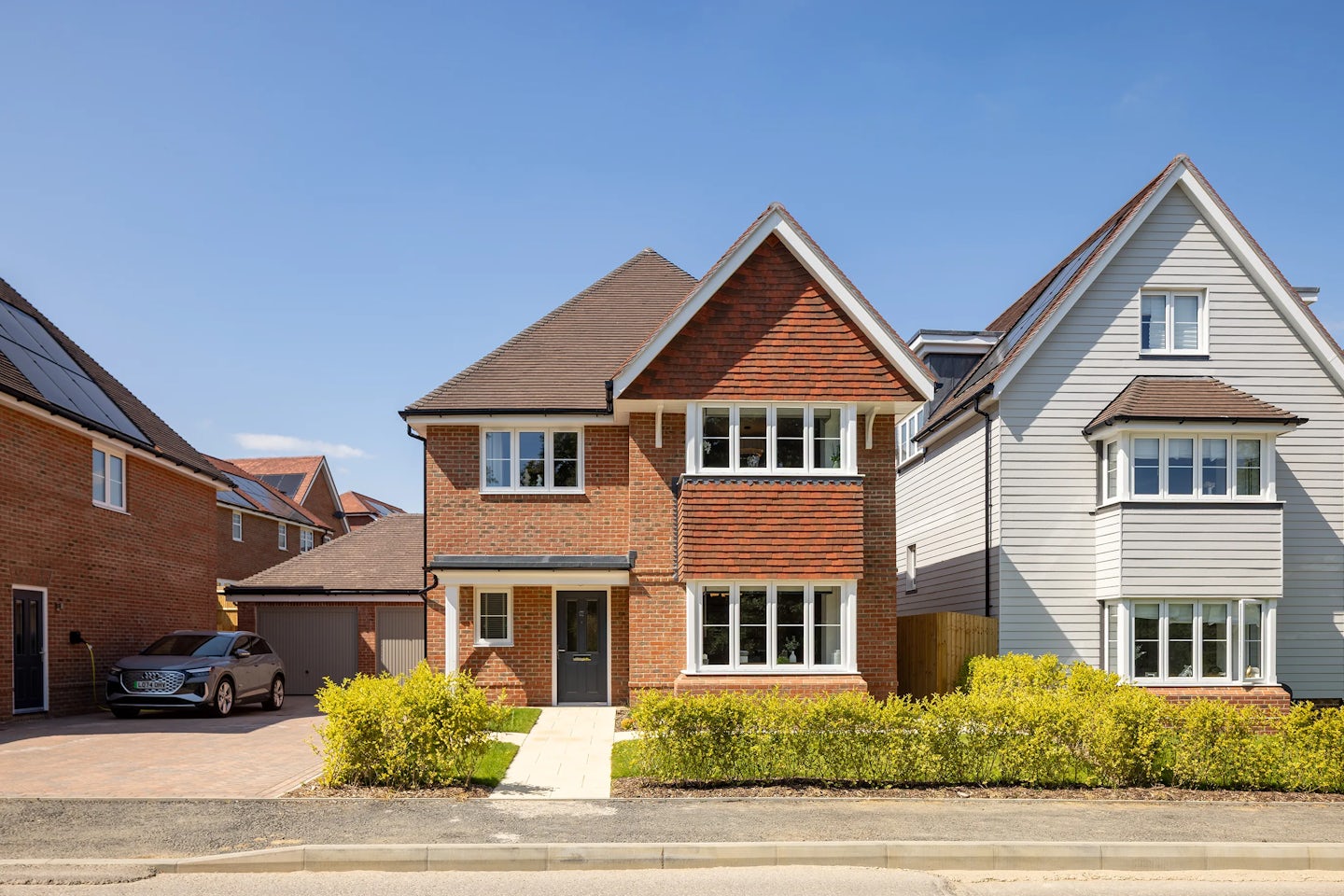A two-story suburban house with a red brick exterior, pitched roof, and multiple white-framed windows, including a prominent bay window on the ground floor. The home sits beside a paved driveway leading to a garage, with a parked car partially visible. Flanked by neighboring houses with contrasting facades—one white and one brick—the scene features a neatly landscaped front yard with shrubs and a walkway to the entrance. Bright sunlight and a clear blue sky evoke a cheerful, residential atmosphere.