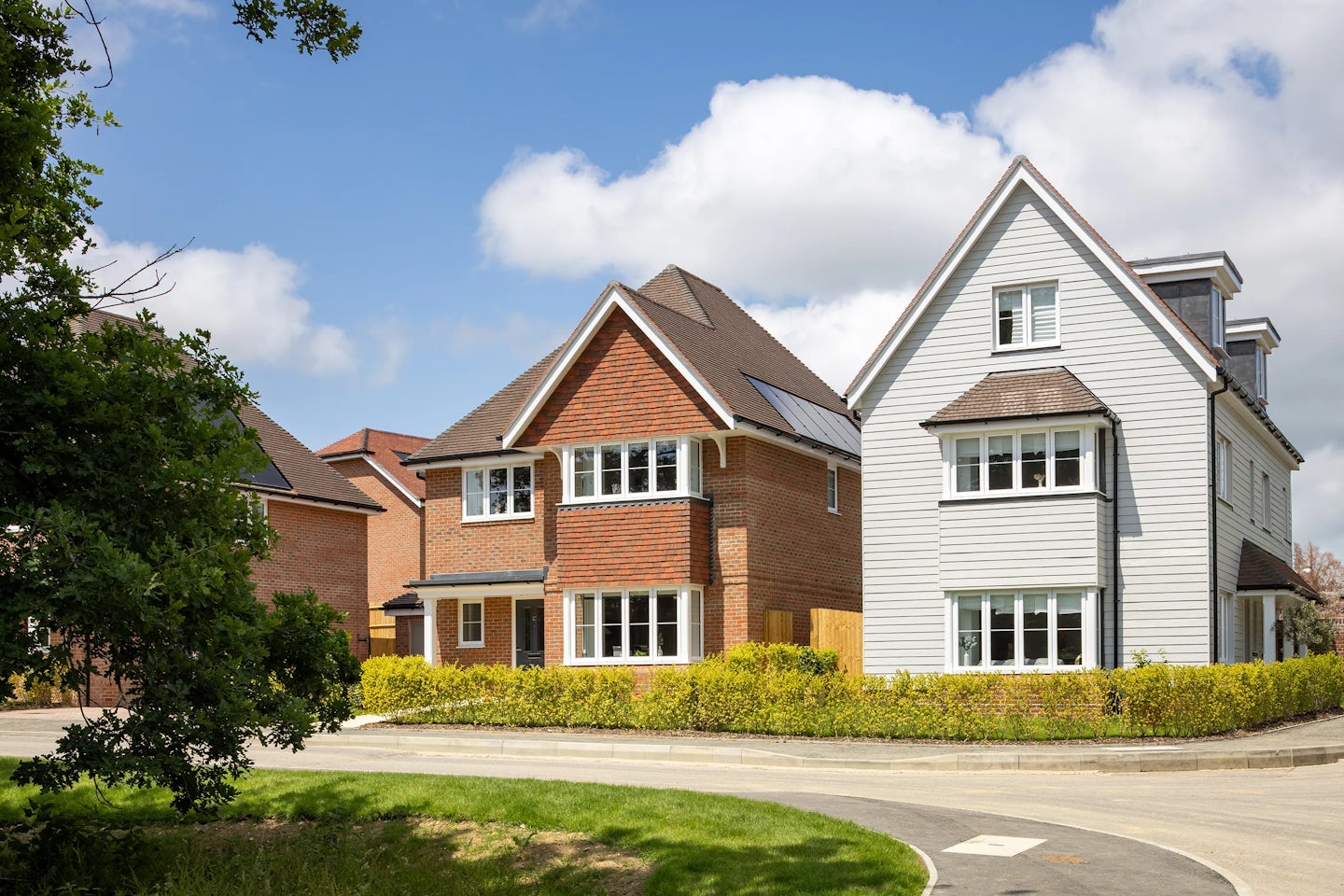 Suburban street view featuring two modern detached homes with pitched roofs and a mix of white siding and red brick exteriors. Both houses have multiple windows and tidy front gardens bordered by hedges. A paved road runs in front, and a tree with leafy branches frames the left edge of the scene. The sky is mostly clear with a few clouds, adding to the peaceful neighborhood atmosphere.