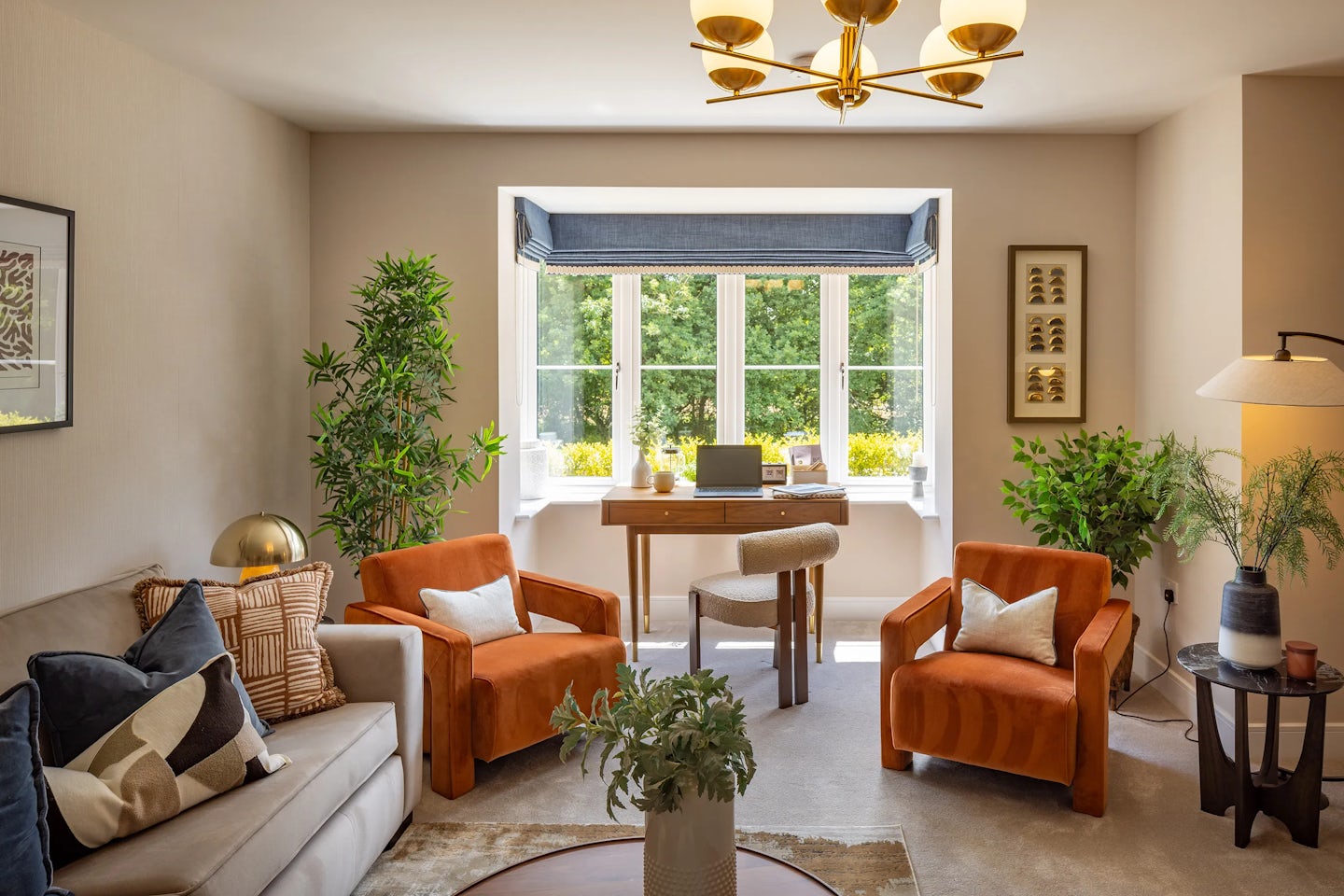 A cozy, eclectic living room filled with natural light from a large window dressed in a soft blue valance. In front of the window, a wooden desk hosts a laptop, globe, and decorative objects, forming a casual workspace. The room features two burnt-orange armchairs with white cushions, a gray sofa with patterned pillows, and a round coffee table with a leafy plant centerpiece. Indoor greenery adds warmth, while framed artwork punctuates the neutral walls. A gold chandelier and a black floor lamp bring layered lighting to the inviting, lived-in space.
