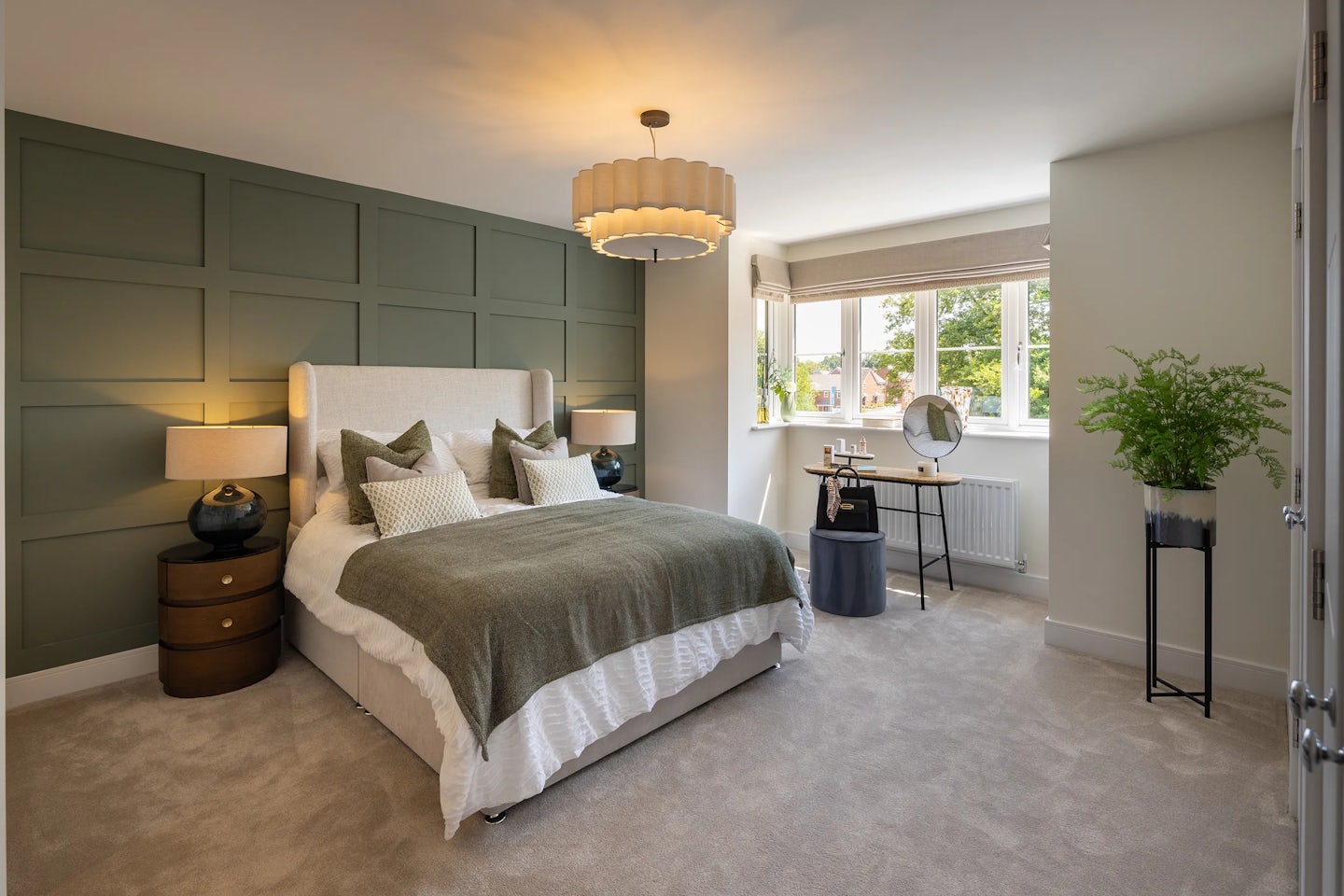 A softly lit bedroom featuring a large bed with a beige upholstered headboard, layered pillows, and a green throw blanket. Twin bedside tables with matching lamps flank the bed, set against a green paneled accent wall. A wide window lets in natural light over a compact desk with a round mirror, white chair, and scattered personal items. A tall leafy plant in a black pot stands beside the desk, adding organic contrast to the beige carpet and warm color palette. A modern ceiling light fixture completes the cozy, balanced ambiance.