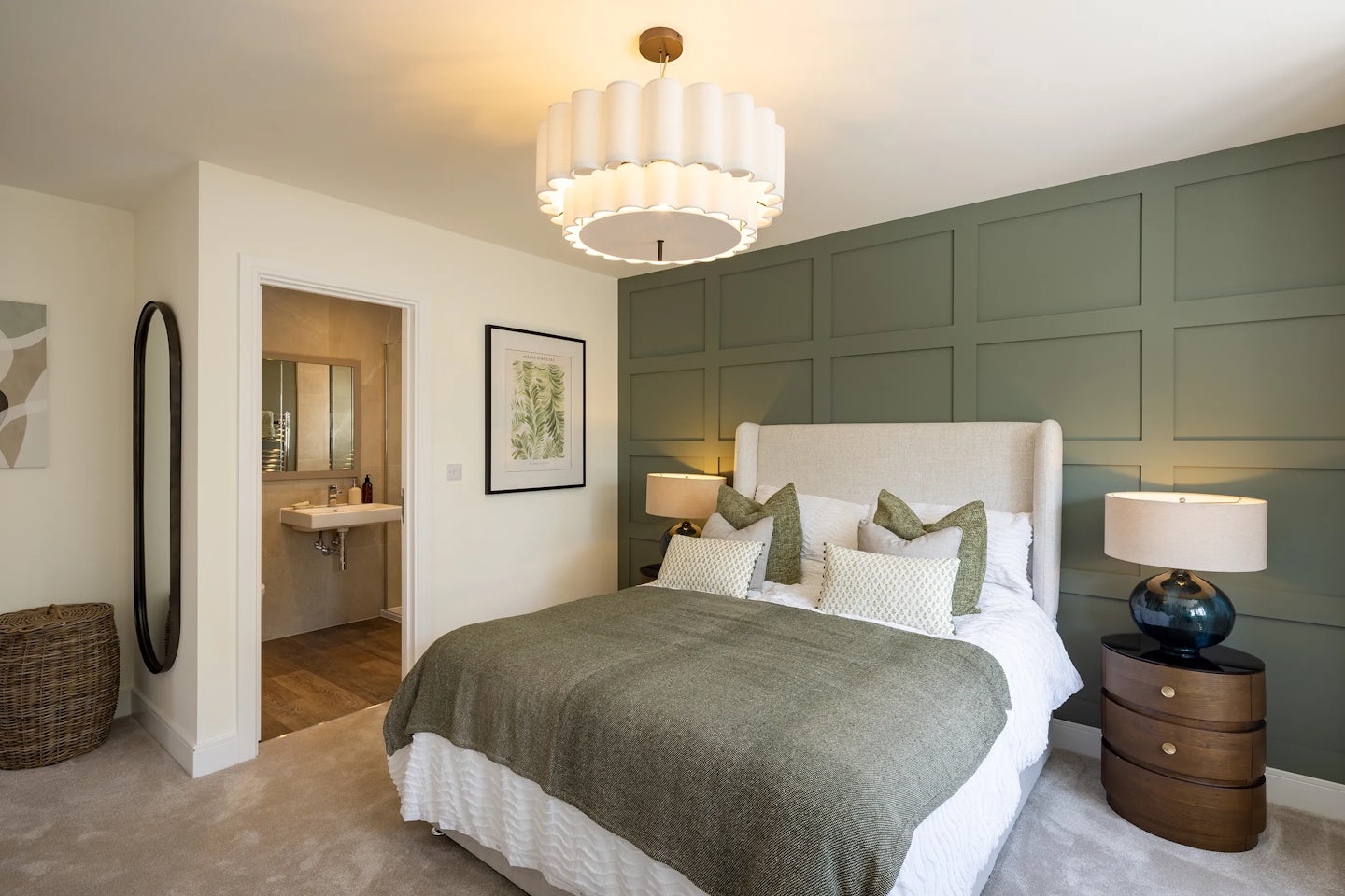 Elegant bedroom with a large beige upholstered bed dressed in green and white pillows and a matching green blanket. A round wooden nightstand with a lamp sits beside the bed, against a dark green paneled accent wall. A statement chandelier with warm lighting hangs overhead, casting a cozy glow. To the left, a doorway leads to a bathroom featuring a sink and mirror. The space includes refined decor: a framed botanical print, a tall oval mirror propped against the wall, and a wicker basket placed nearby
