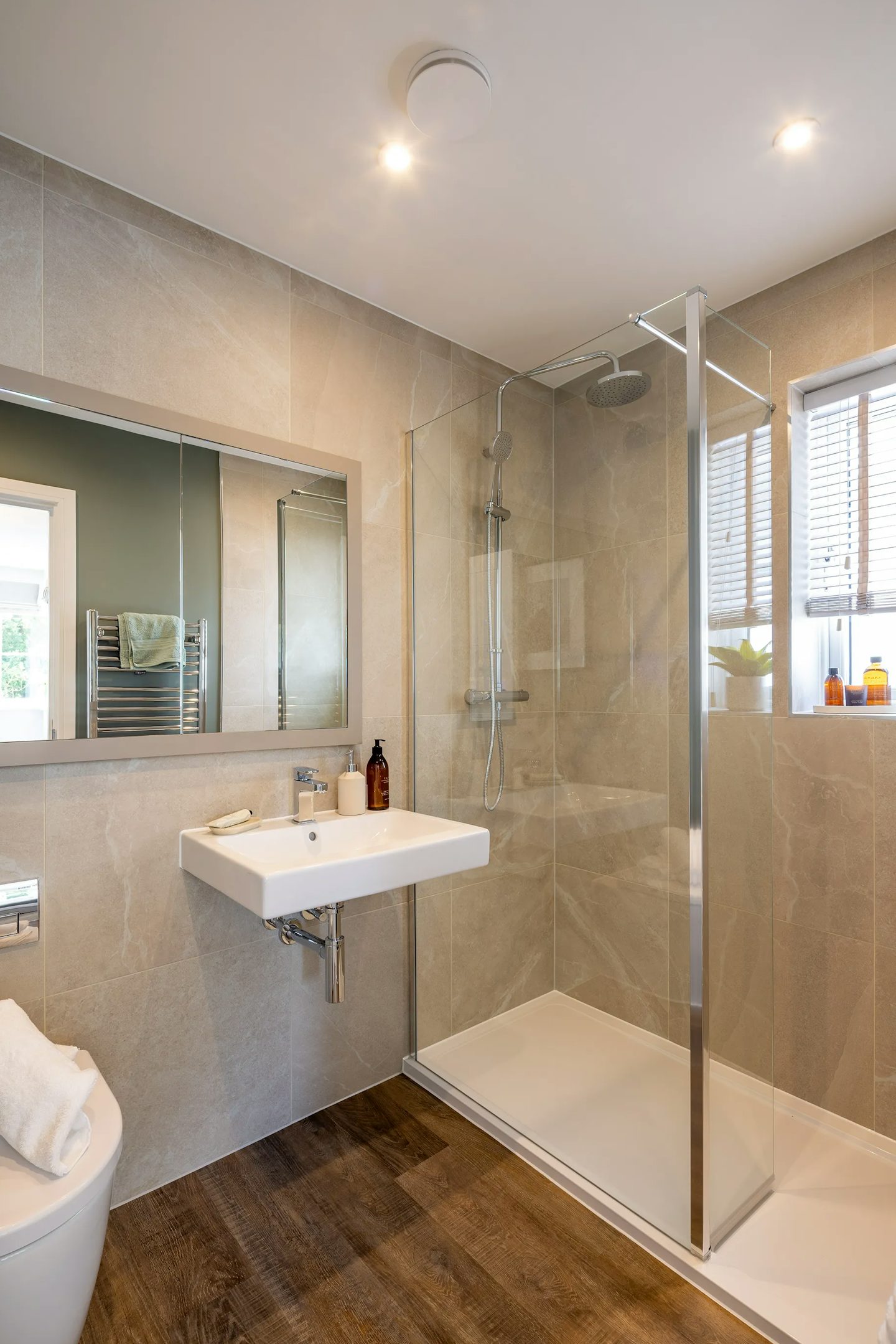 Contemporary bathroom with a glass-enclosed shower featuring both rainfall and handheld showerheads. A white wall-mounted sink with a chrome faucet sits beneath a large mirror, while a toilet with a neatly folded white towel adds a touch of hospitality. Warm wood flooring contrasts with beige tiled walls, and natural light streams in through a window fitted with blinds.