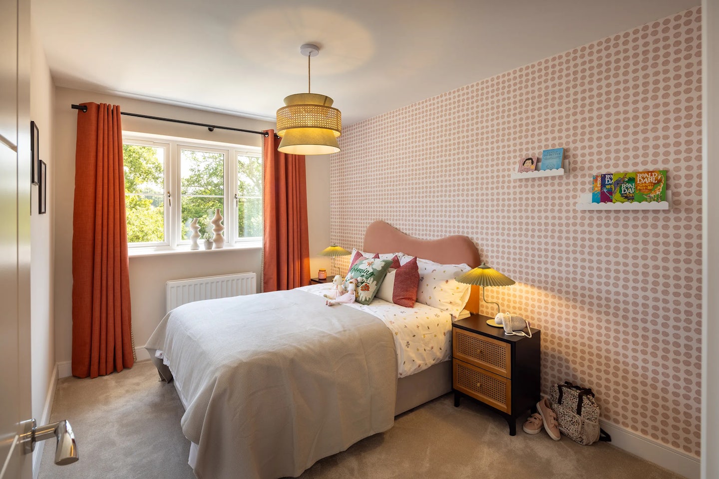 A cozy, thoughtfully styled bedroom with a double bed dressed in a light grey duvet and patterned white sheets. A mix of pillows—brown, beige, and subtly textured—adds depth to the bedding. The backdrop features circular-pattern wallpaper in muted tones, framing twin bedside tables topped with yellow-shaded lamps. Above the bed, two compact shelves display framed artwork, colorful books, and small decorative items.