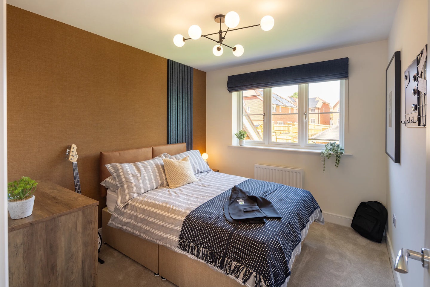 A stylish bedroom with warm lighting and a cozy, personal feel. The bed is dressed in striped bedding with layered pillows, a black jacket and sunglasses casually placed on top. A sculptural chandelier hangs overhead, adding a touch of elegance. To the left, a wooden bedside table supports a leafy plant, while a guitar leans against the textured brown accent wall beside it. A black vertical panel adds contrast behind the bed. On the right, a window with dark blue blinds reveals neighboring houses and sits above a white radiator.