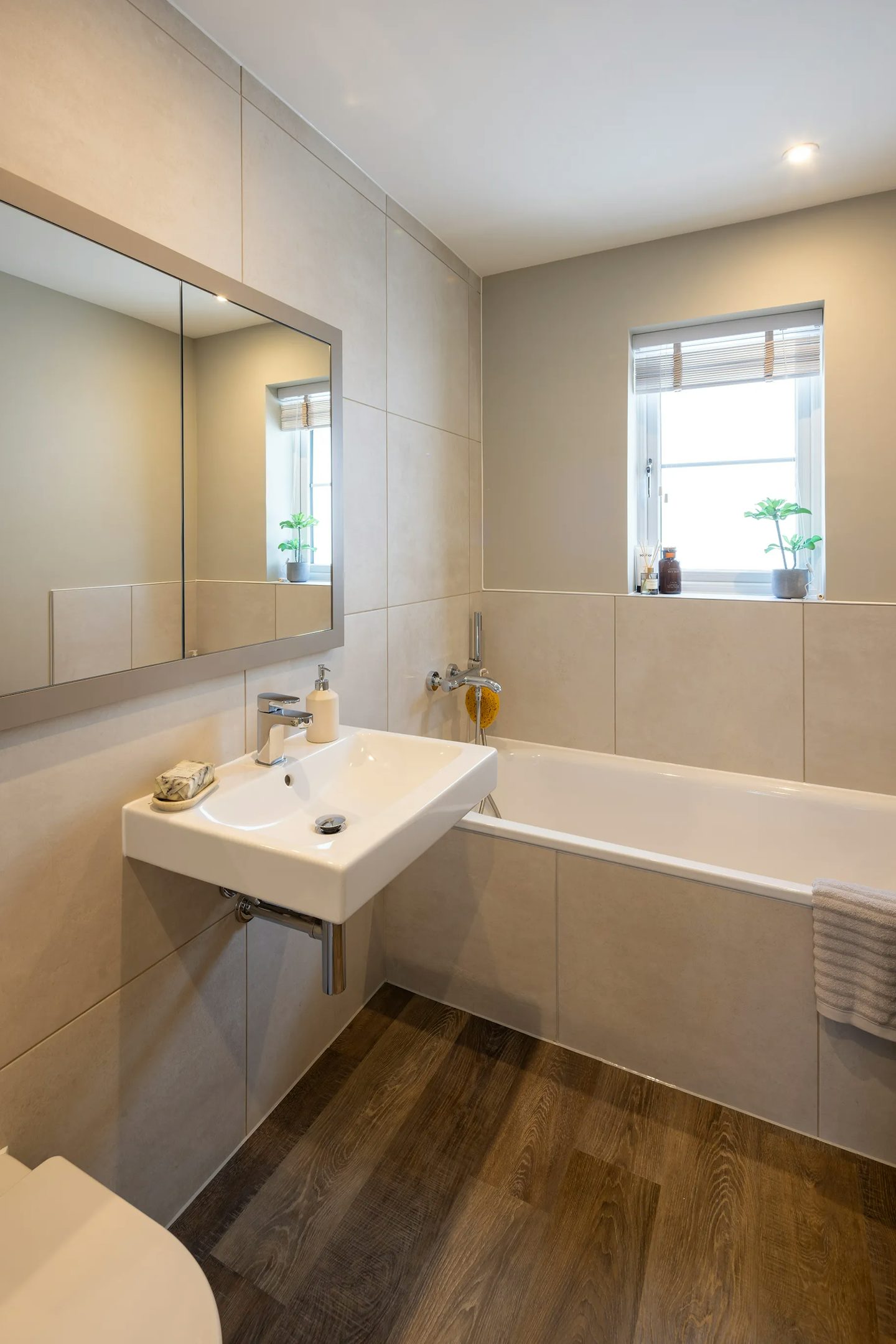A bright, modern bathroom with a serene aesthetic. The space features a white basin with a chrome tap, flanked by a soap dispenser and a bar of soap. A large wall mirror reflects the clean lines of the room. In the background, a white bathtub is partially visible with a folded towel draped over its edge. A window above the tub lets in natural light and showcases two small potted plants on the windowsill.