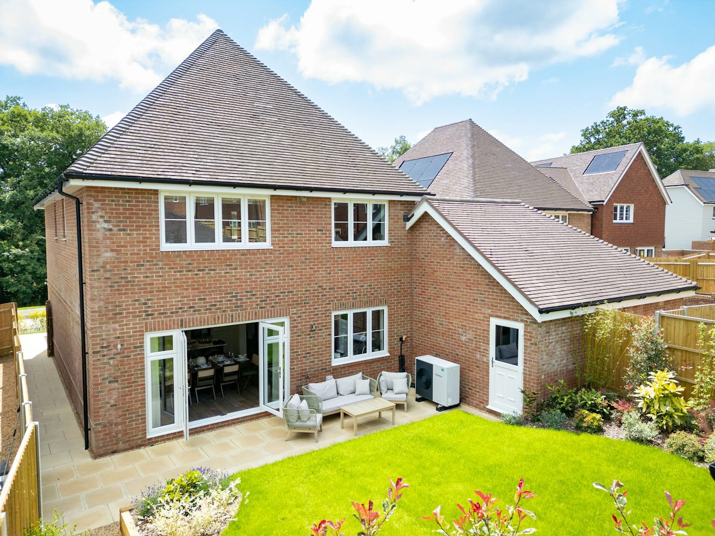 A modern two-story brick home with a sloped roof and multiple white-framed windows, set behind a well-kept backyard. The garden includes a paved patio with rattan-style furniture—a sofa, chairs, and a table—arranged for outdoor lounging. Decorative plants border the space, and a white door connects the patio to the house. Neighboring homes are visible, one featuring solar panels on the roof, hinting at eco-conscious living. The scene is bathed in sunlight beneath a partly cloudy sky, offering a bright, suburban atmosphere.