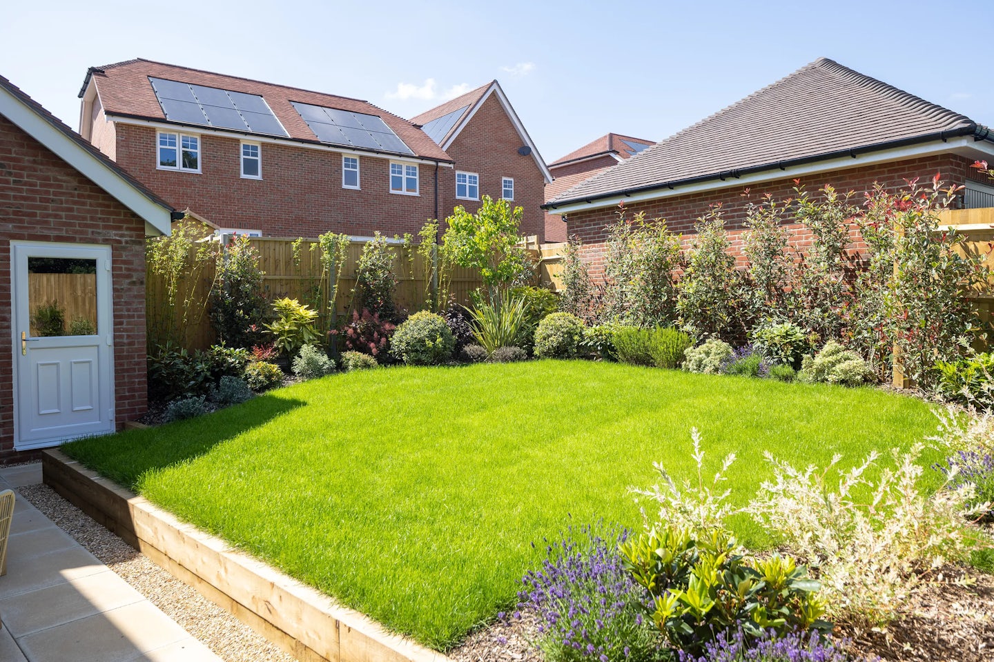 Lush backyard garden featuring a vibrant green lawn bordered by dense shrubs and climbing plants. A small paved patio area sits to one side, partially shaded by nearby foliage. In the background, two brick houses with pitched roofs are visible, each equipped with rooftop solar panels—suggesting an eco-friendly touch. The garden is enclosed by a wooden fence, and bright sunlight casts soft shadows across the well-tended space, evoking a peaceful suburban atmosphere.