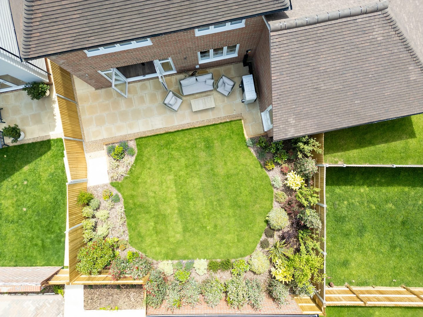Aerial view of a landscaped backyard garden featuring a lush green lawn bordered by flower beds and leafy shrubs. A paved patio area sits adjacent to the house, furnished with a gray outdoor sofa, matching chairs, and a low table, creating a relaxed seating arrangement. The garden is enclosed by wooden fencing, and a stone pathway connects the patio to the lawn. Trees and climbing plants add privacy and natural charm to the peaceful outdoor space.