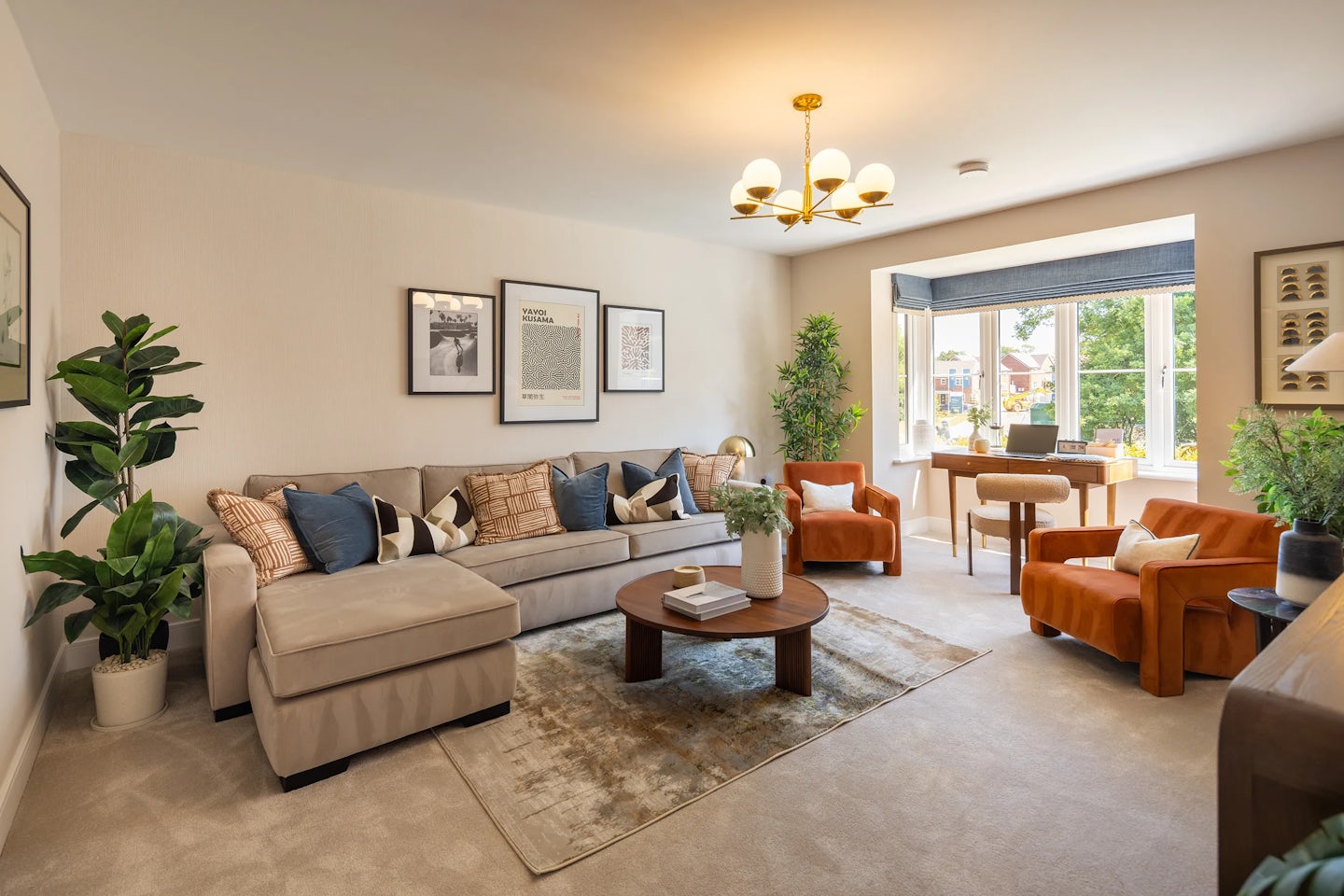 Warm and inviting living room featuring a large beige sectional sofa adorned with assorted patterned cushions. Above the sofa hang three framed artworks, adding subtle visual interest. A round wooden coffee table sits at the center, styled with a leafy plant and stacked books. To the side, two burnt-orange armchairs face a wide window that floods the space with natural light. A wooden desk and chair form a compact workspace by the window, while potted plants and a six-light chandelier enhance the room’s cozy, lived-in charm. Beige carpeting ties the space together with a neutral, grounding texture.