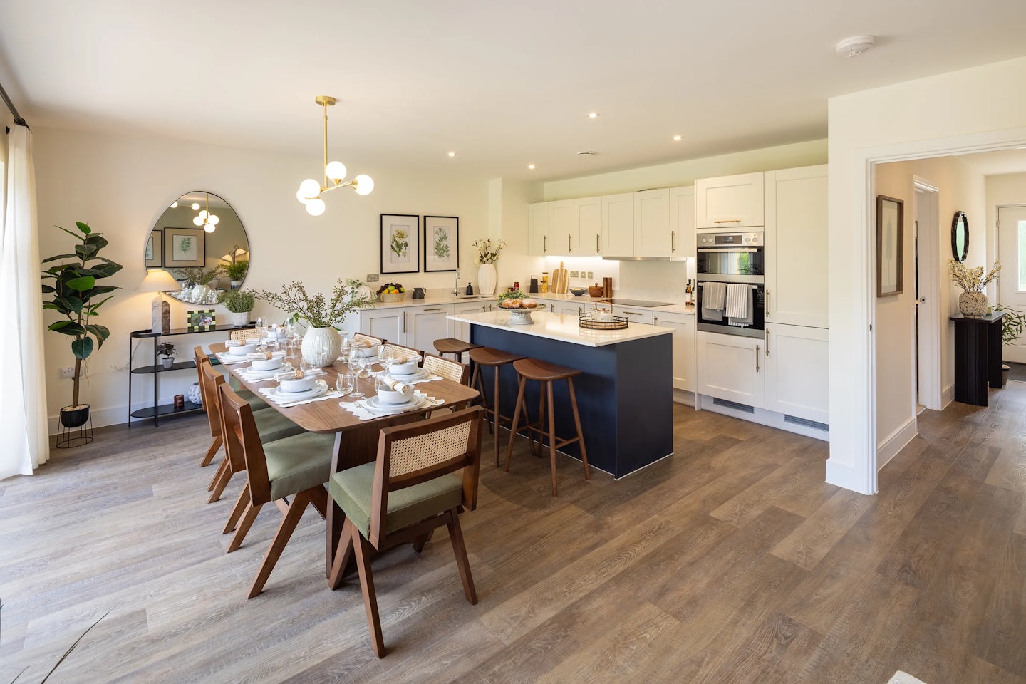 Stylish open-plan kitchen and dining area with a wooden table set for six, each place arranged with white plates, glasses, and silver cutlery. A floral centerpiece adds a touch of elegance beneath a spherical chandelier. Behind the table, a modern kitchen features white cabinetry, stainless steel appliances, and a dark island with two wooden bar stools. The space includes decorative wall elements like framed artwork, a round mirror, and potted plants. Warm wooden flooring and ambient lighting create a welcoming, polished atmosphere.