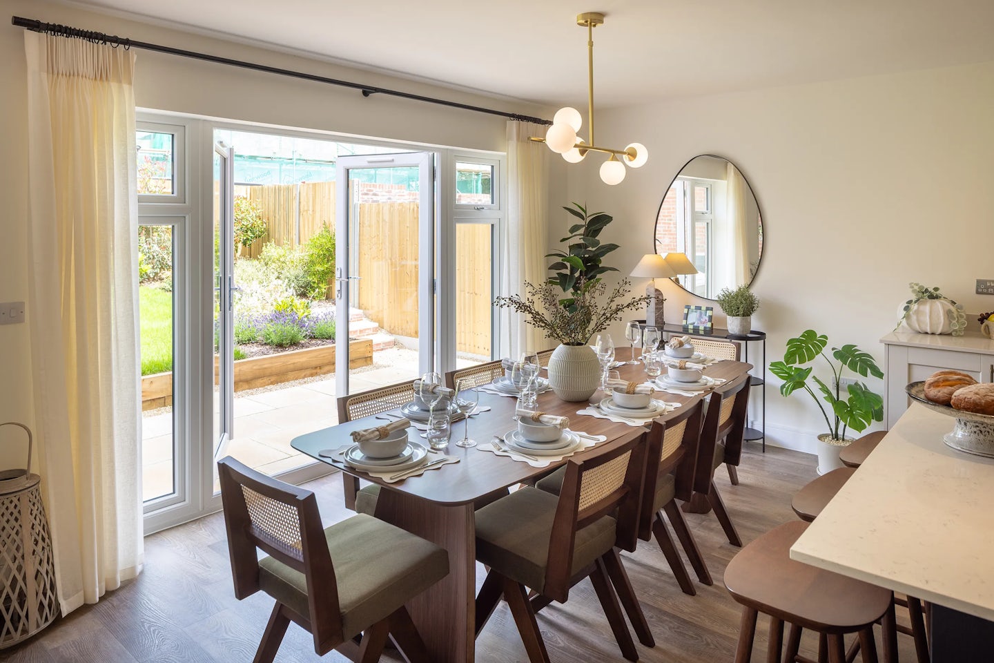 Elegant dining room with a long wooden table set for eight, featuring white plates, bowls, glasses, and cutlery arranged neatly. A glass vase with fresh flowers and greenery serves as a centerpiece beneath a modern chandelier with spherical lights. The space is framed by large glass doors opening to a patio and garden, allowing ample natural light to flood the room. On the right wall, a round mirror, decorative plants, and a lamp contribute to the room’s inviting and refined aesthetic. Would you like to shift this toward a tone that emphasizes emotional warmth, sustainability features like natural light, or align it with a specific platform? I’m happy to refine it with you.
