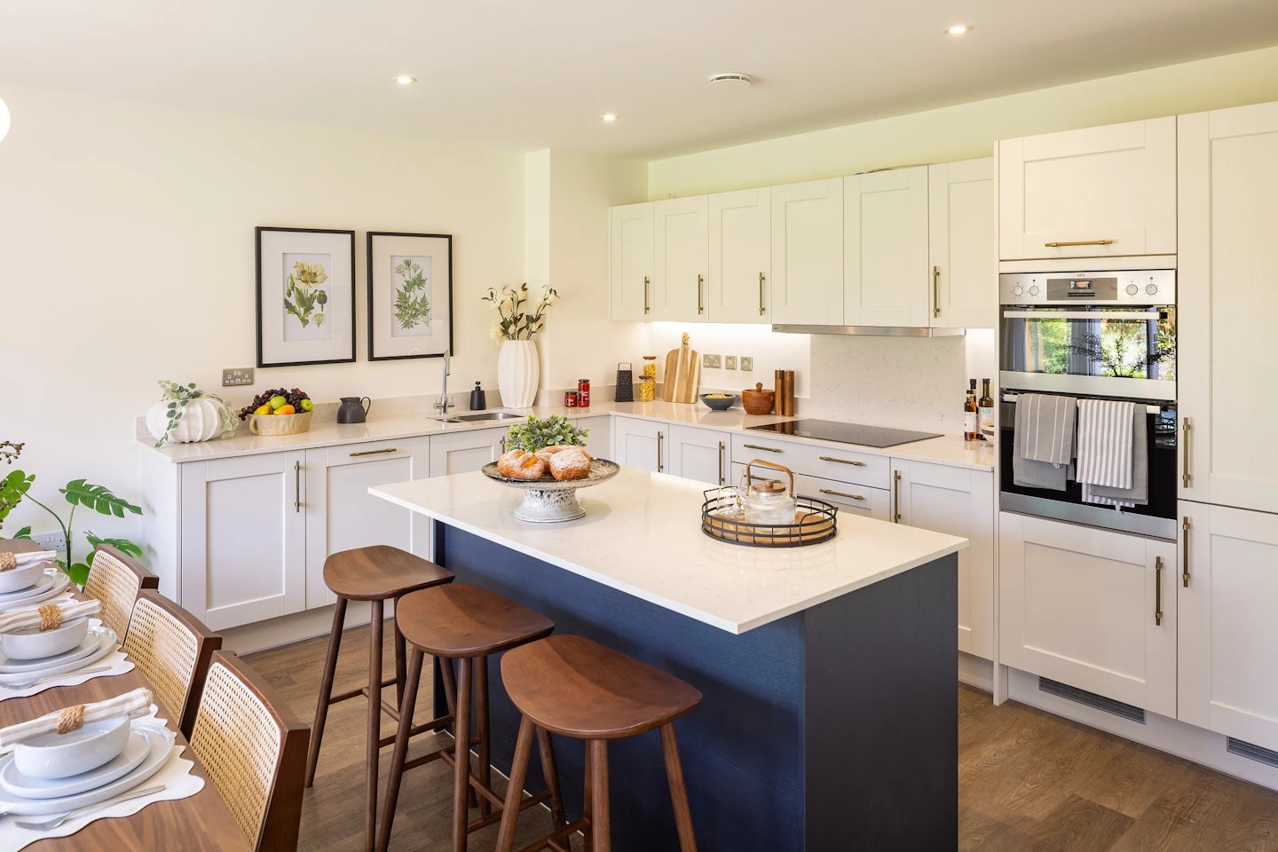 A spacious, light-filled kitchen with a central island featuring a white countertop and deep blue base, paired with three wooden bar stools. The island displays a cake stand with pastries and a wire basket with a lid, adding a touch of everyday charm. Surrounding white cabinets with gold handles contrast elegantly with the darker tones, while a built-in oven and stovetop anchor the cooking space. A nearby countertop holds a mix of decorative and functional items including a vase of flowers, a pumpkin, and framed botanical prints above. In the foreground, a wooden dining table is neatly set with plates, bowls, and napkins, accompanied by a leafy plant in the corner for a fresh, welcoming touch.