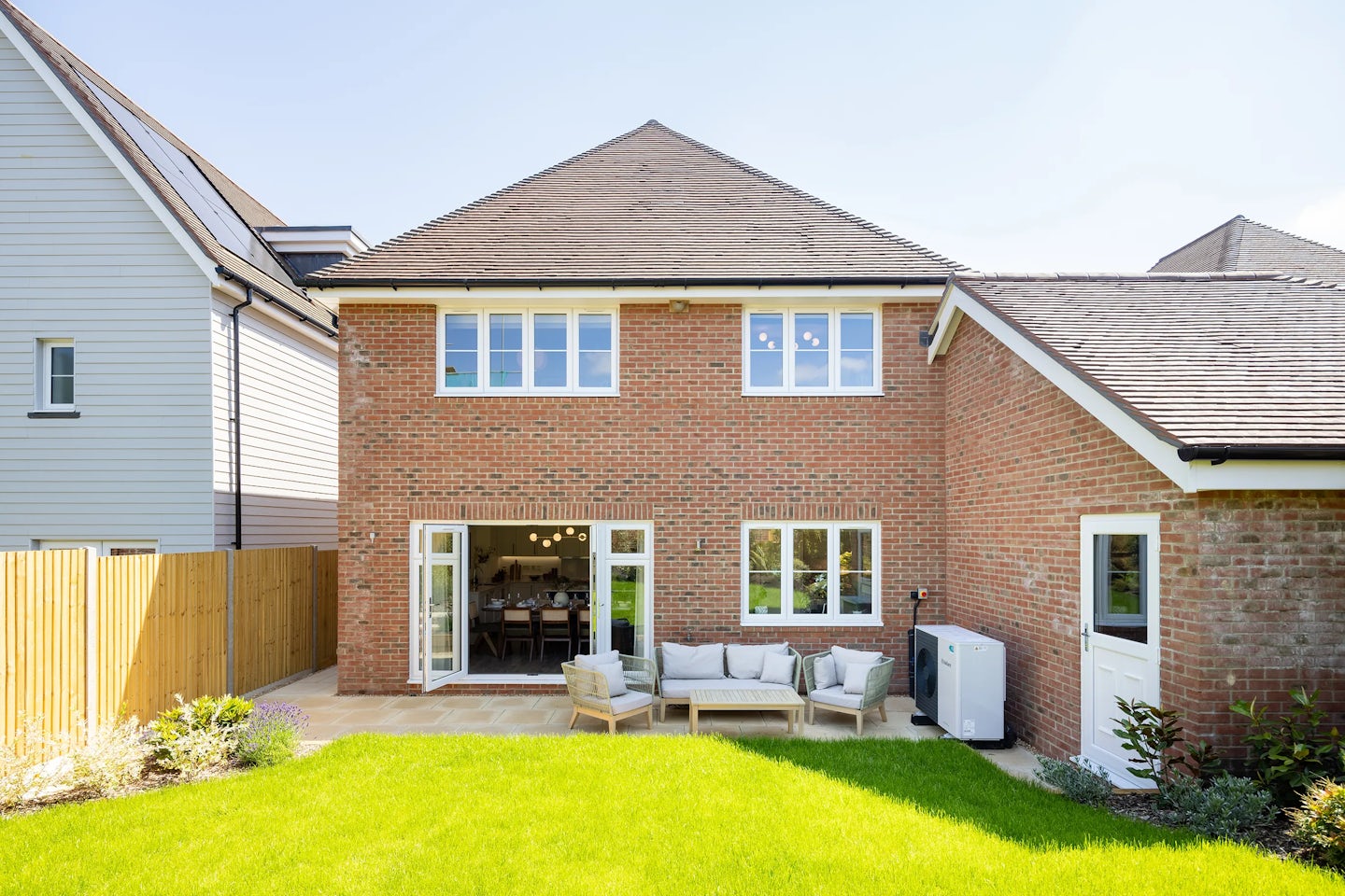 Suburban backyard featuring a well-maintained lawn bordered by neat planting beds and a paved patio area with outdoor seating, including a modern rattan-style sofa and matching chairs. The house has a red brick exterior, pitched roof, and large white-framed windows, with an air conditioning unit visible on the side. Neighboring homes and fencing frame the scene, while bright sunlight casts crisp shadows, creating a fresh and welcoming residential atmosphere.