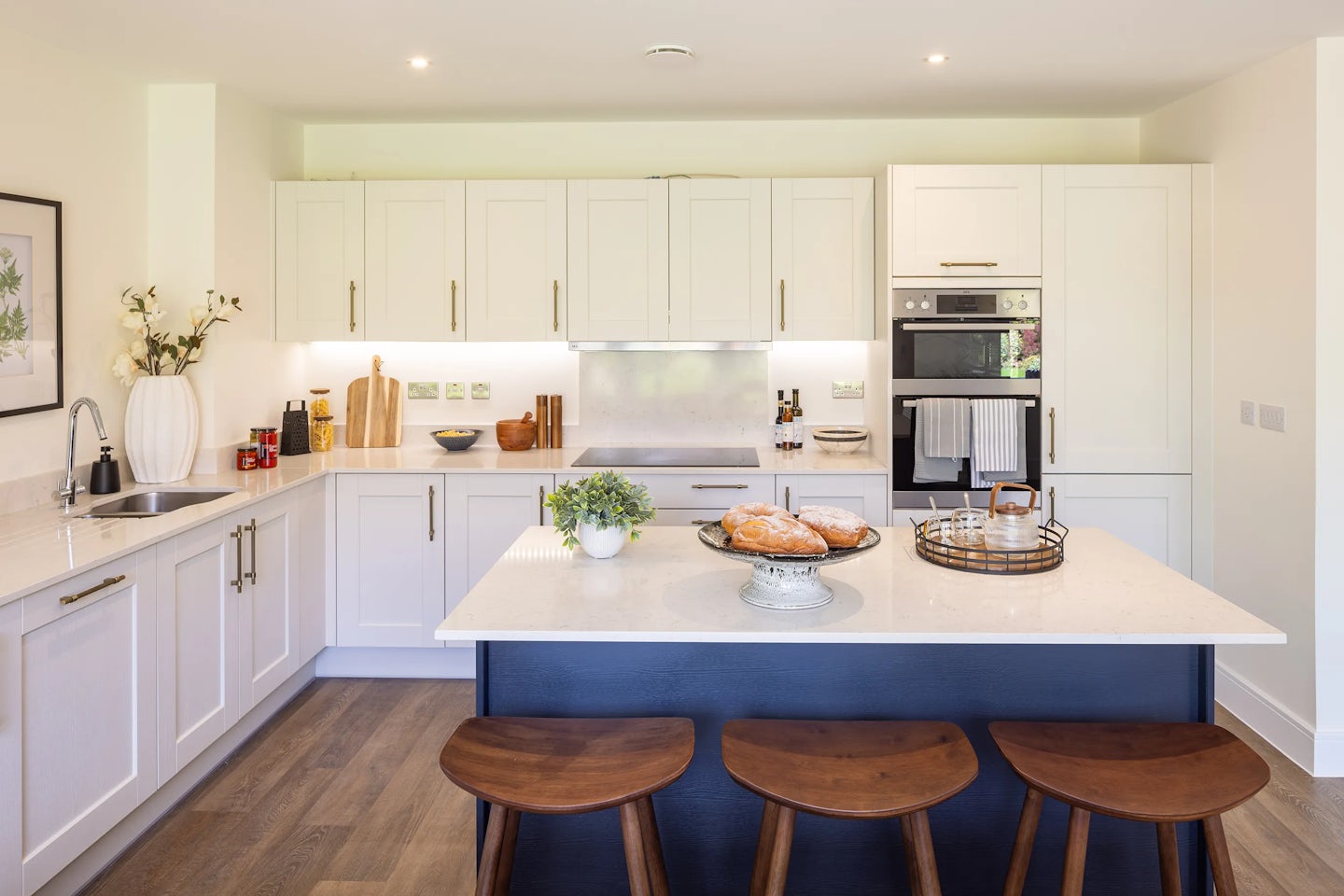Modern kitchen featuring white cabinetry, built-in oven and microwave, and a central island with a smooth white countertop and wooden base. The island is styled with a white potted plant, a loaf of bread on a plate, and a woven basket with a cloth liner, suggesting warmth and everyday functionality. Three wooden stools tuck neatly under the island, while the surrounding space includes cutting boards, storage jars, and a vase of flowers that add personality and charm. Wooden flooring and bright lighting enhance the clean, inviting atmosphere.