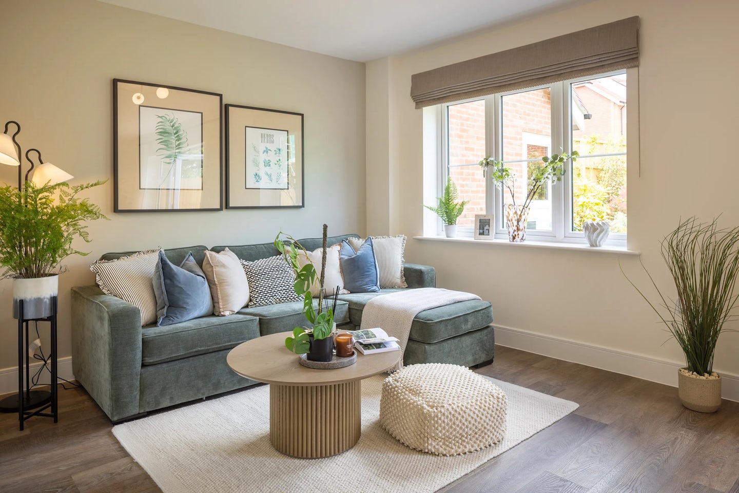 Bright and inviting living room centered around a green sectional sofa adorned with mix-toned pillows in white, blue, and gray. A round wooden coffee table sits atop a light rug, styled with candles, a plant, and stacked books, while a textured pouf adds casual comfort. The space includes several indoor plants—one on a black metal stand beside the sofa and others lining the sunny windowsill—creating a lively, organic feel. Two framed botanical prints accent the wall, and the soft natural light highlights the warm, relaxed atmosphere.