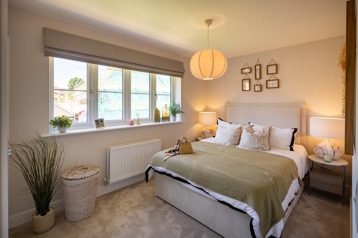 Thoughtfully styled bedroom featuring a double bed dressed in crisp white bedding and a green throw blanket, accented with coordinating pillows. Two bedside tables hold matching lamps and small decorative objects, while above the bed, a gallery-style arrangement of framed artworks creates a focal point. A large window with a beige blind lets in natural light and offers a partial view of a construction site outside. Nearby, a potted plant and a wicker basket rest on the beige carpet, adding warmth and texture. A modern ceiling light enhances the calm, curated ambiance.