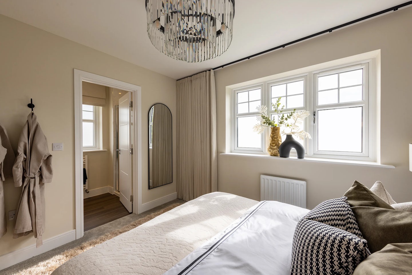 Elegant, light-filled bedroom with a large window dressed in sheer white curtains, allowing sunlight to stream into the space. On the windowsill sit a black abstract sculpture, a vase of yellow flowers, and a small plant, adding personality and texture. The bed is neatly made with white bedding and patterned accent pillows. To the left, a doorway opens to an adjoining room, and a full-length mirror is mounted beside it. A white robe hangs on a wall hook, and a modern chandelier with globe lights provides ambient lighting from above.