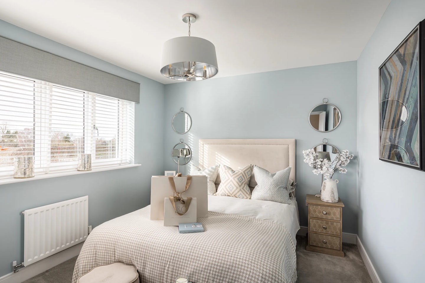 Tastefully styled bedroom with a soft blue accent wall and beige upholstered headboard framing a neatly made bed. The bed features a beige duvet, patterned pillows, and a shopping bag and book placed casually on top, suggesting a lived-in feel. A round mirror and wall lamp hang above the bed, while a bedside table holds a small vase of white flowers and a book. Natural light filters through white blinds beneath a grey valance, adding to the serene ambiance. A modern ceiling light and abstract framed artwork on the right wall complete the calming, cohesive design.