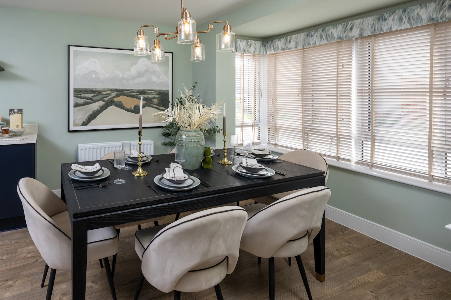 Modern dining room featuring a rectangular black dining table surrounded by six beige upholstered chairs. The table is neatly set with white plates, glasses, cutlery, and decorative accents including a vase of dried flowers, two candlesticks, and a small potted plant. Overhead, a contemporary chandelier with five glass pendant lights adds a stylish focal point. Large windows with wooden blinds let natural light pour into the space