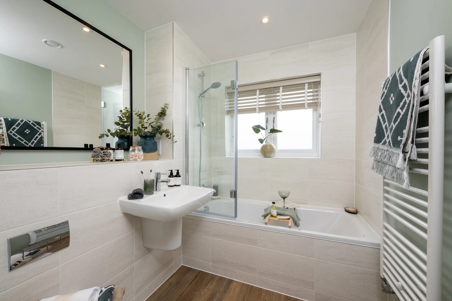 Contemporary bathroom featuring a white sink with chrome fixtures set beneath a large rectangular mirror. A bathtub with a glass shower screen sits beside a window with horizontal blinds, allowing natural light to brighten the space. Decorative touches include potted plants, soap dispensers, and neatly folded towels—one hanging from a wall rack with a geometric pattern.