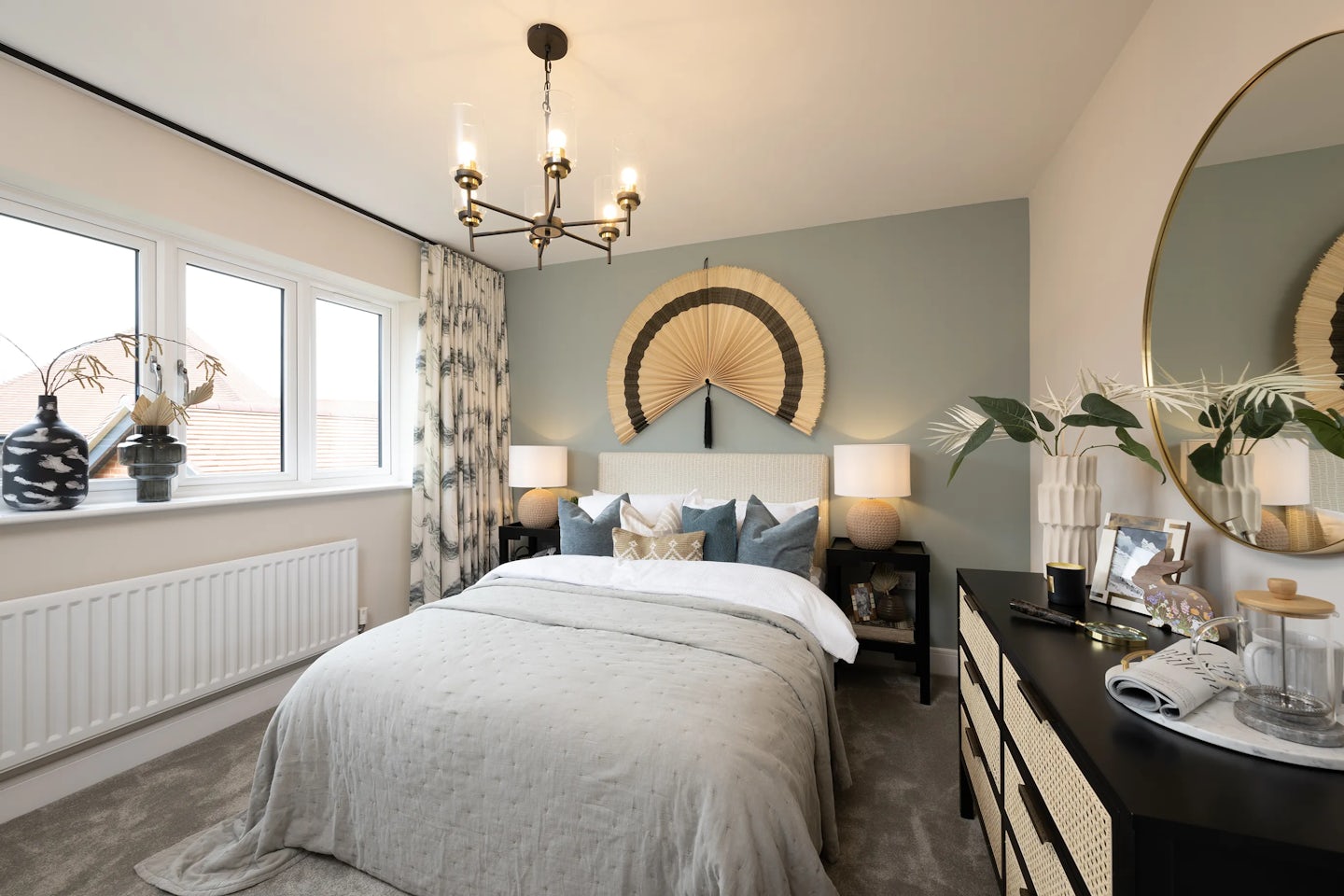 Stylish, softly lit bedroom featuring a double bed with a grey bedspread and assorted pillows in muted tones. Above the bed hangs a fan-shaped decorative wall piece that adds texture and visual interest. Matching nightstands flank the bed, each topped with a warm-glow lamp. To the left, a window with patterned curtains and a white radiator below allows daylight into the room. On the right, a dark dresser with woven drawer fronts holds plants, a framed picture, and assorted personal items, while a circular mirror is mounted above.