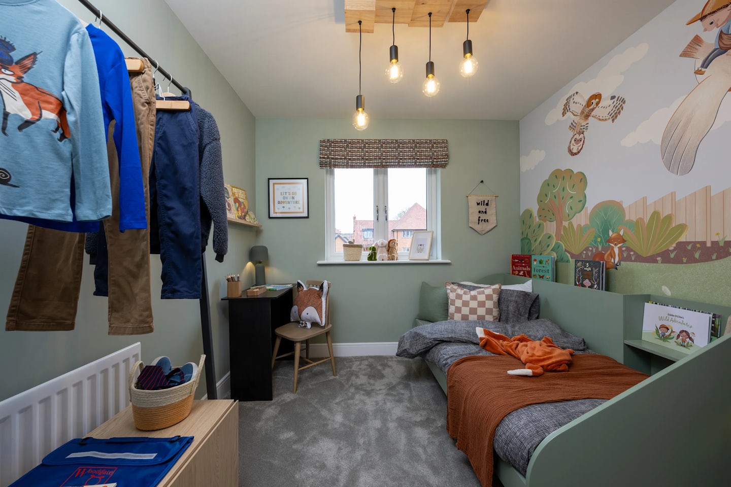 Whimsically styled children’s bedroom with a cozy layout and gentle natural light. On the left, a small wooden table sits beside a basket of toys and a clothes rail displaying neatly hung garments including a blue fox-print shirt and brown trousers. A patterned blind shades a central window, below which a desk holds books, framed art, a lamp, and a potted plant. To the right, a single bed with a grey duvet and orange throw nestles under a soft-toned wall mural featuring birds and trees, adding playful charm.